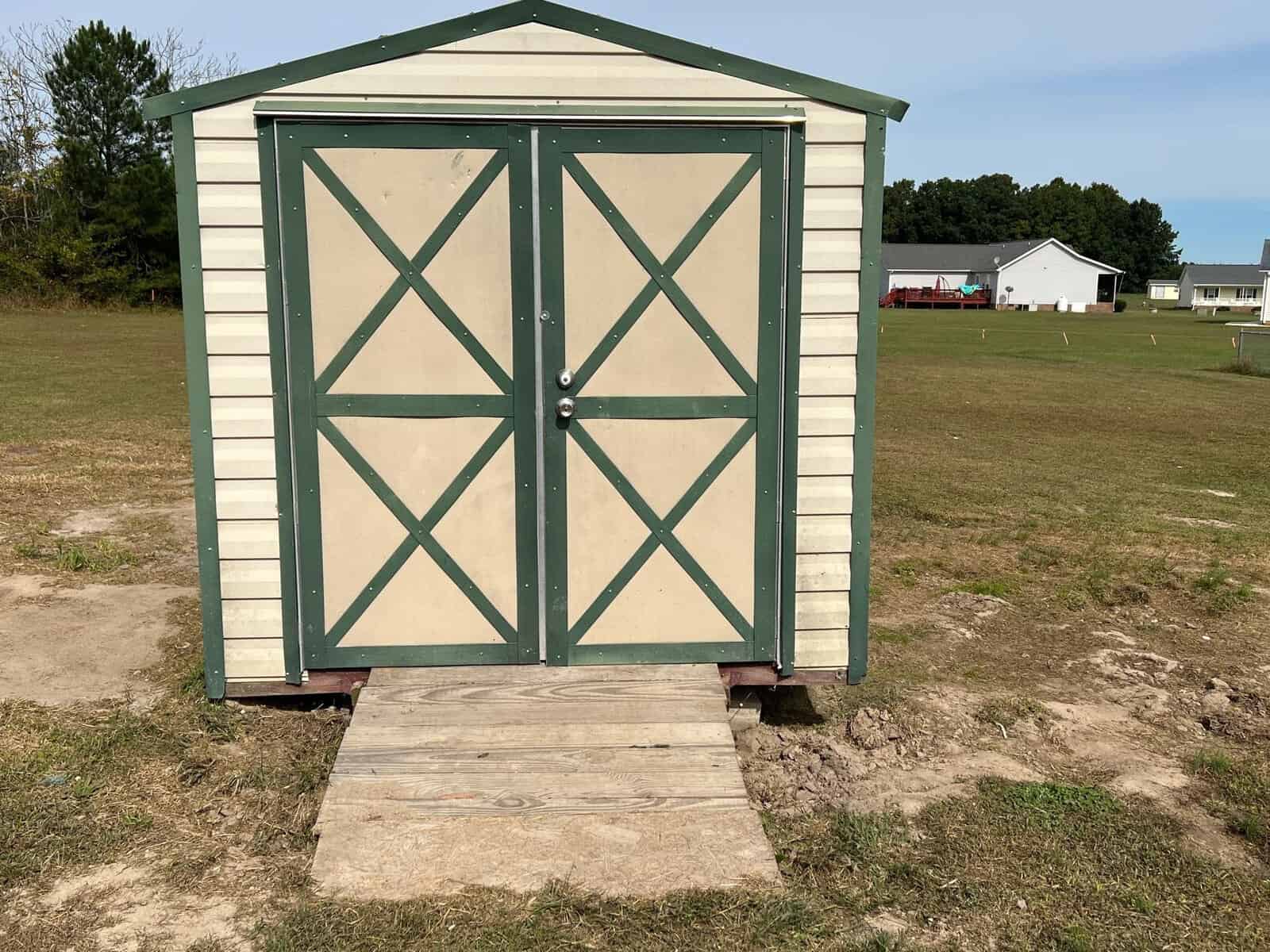 A small beige storage shed with green trim and double doors stands on a grassy area with a simple wooden ramp leading to the entrance.