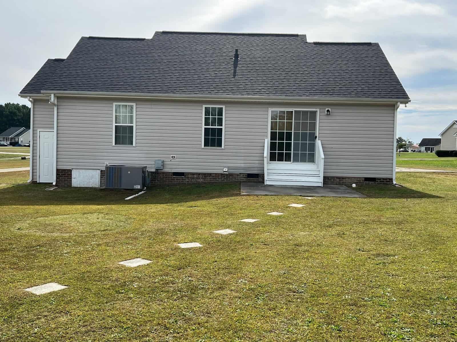 A suburban single-story house with beige siding features a white door, two windows, an external hvac unit, and a series of stepping stones leading to the backyard on a clear day.