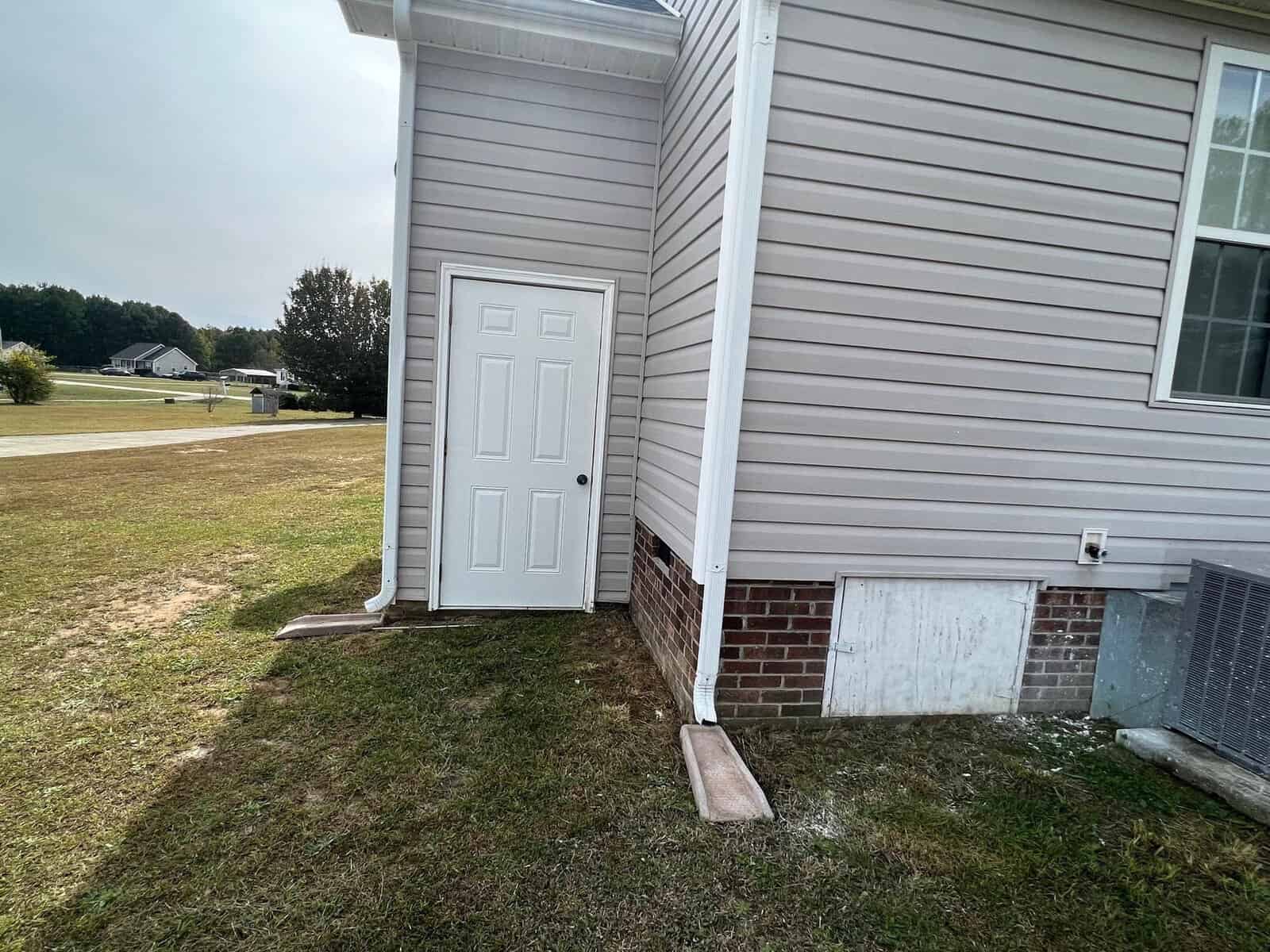 A side door of a house with vinyl siding, a small brick exterior at the base, an air conditioning unit to the right, and a wooden plank lying on the ground in front of the door.