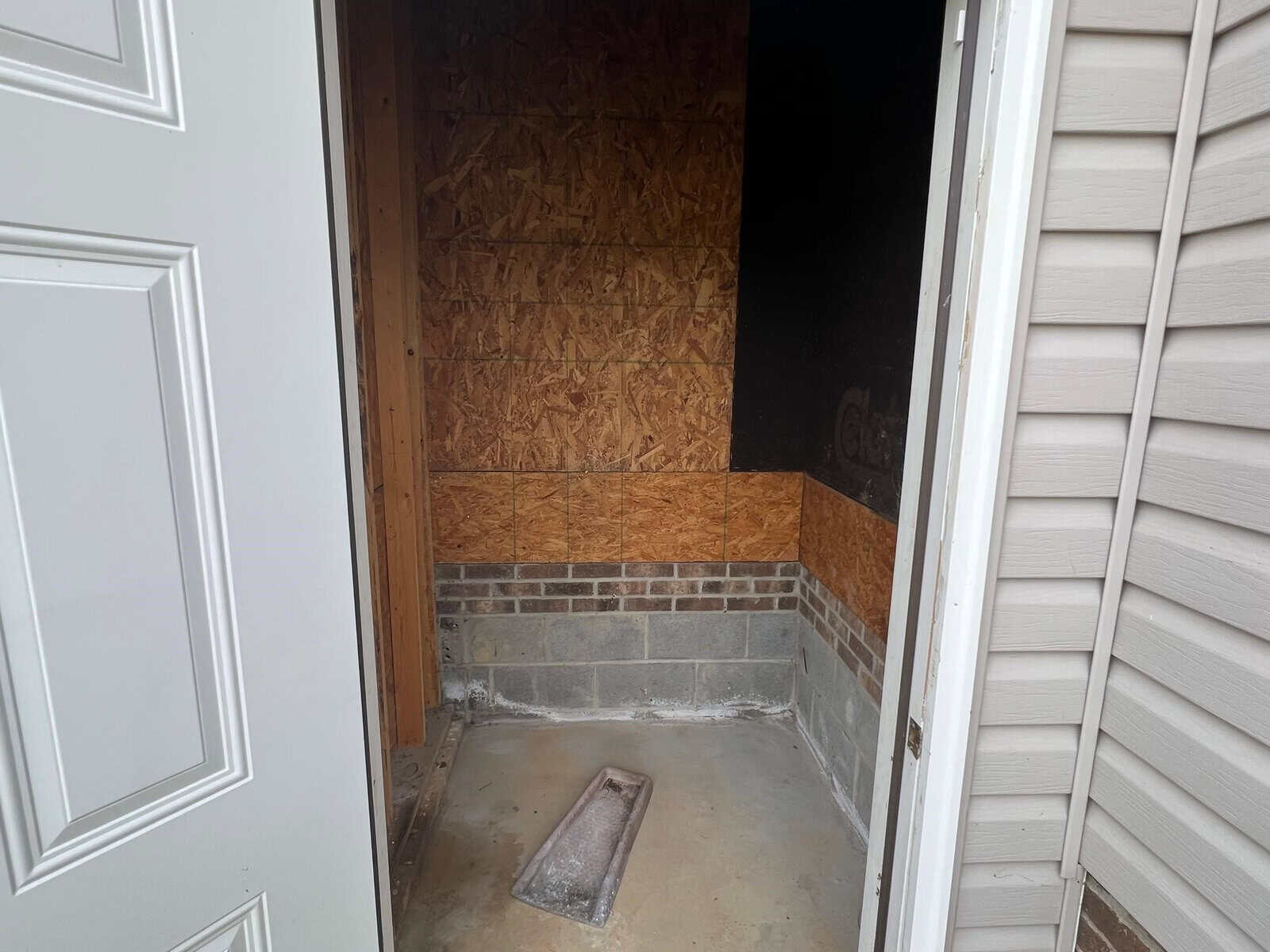 An unfinished storage area or small room with exposed plywood walls, a concrete floor, and a single step leading inside from the doorway.