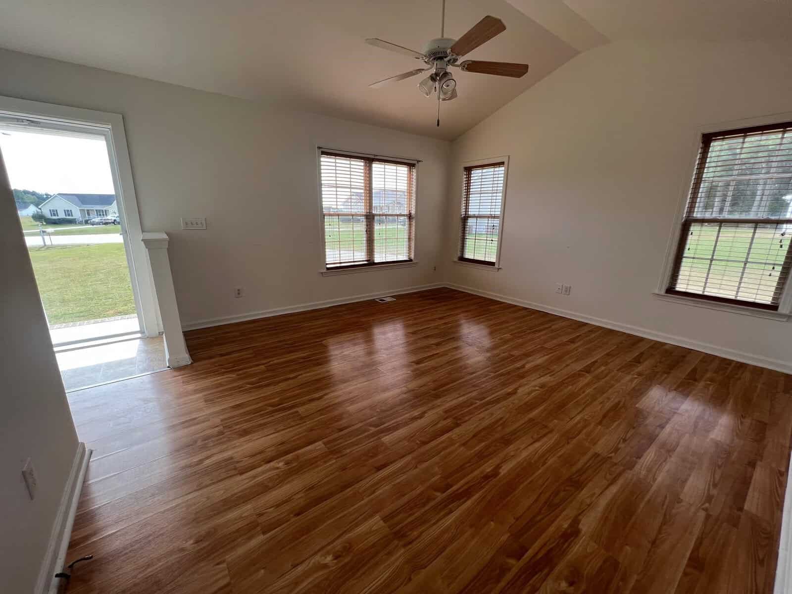An empty room with polished wooden flooring, a ceiling fan overhead, and two windows providing a view of the outside.