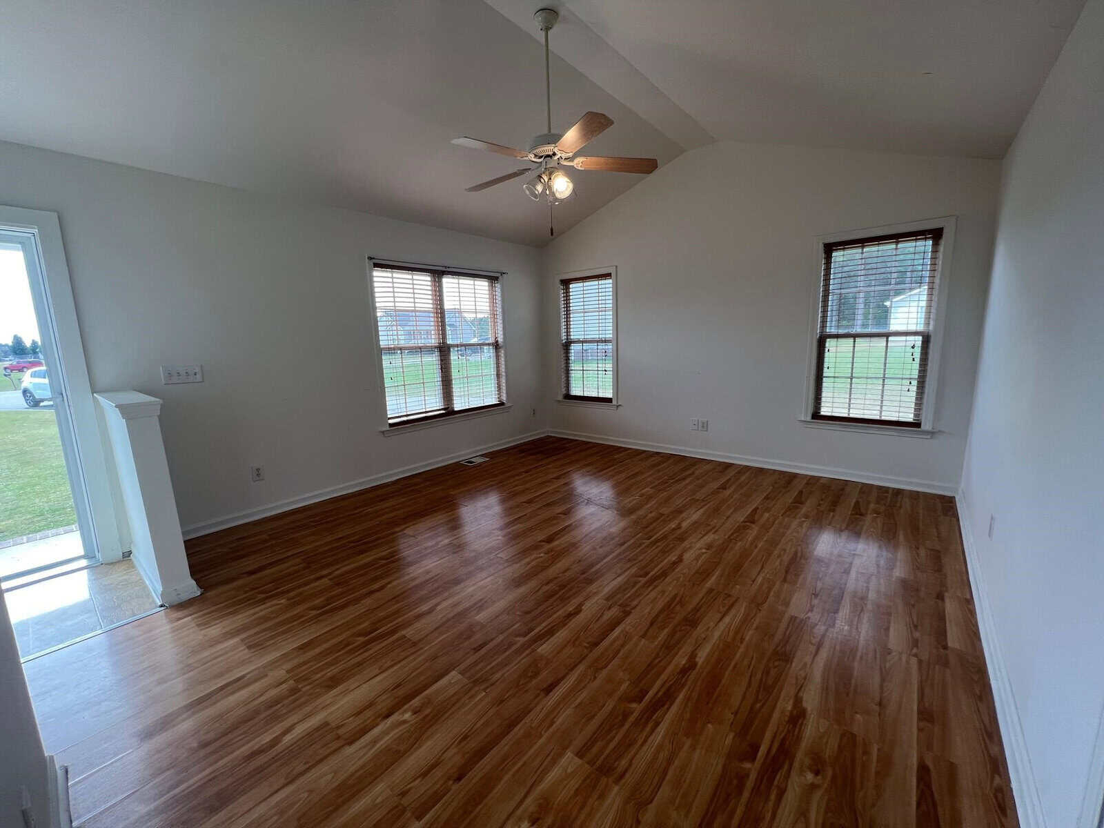 An empty room with wood laminate flooring, white walls, and a ceiling fan, featuring three windows that provide natural light.
