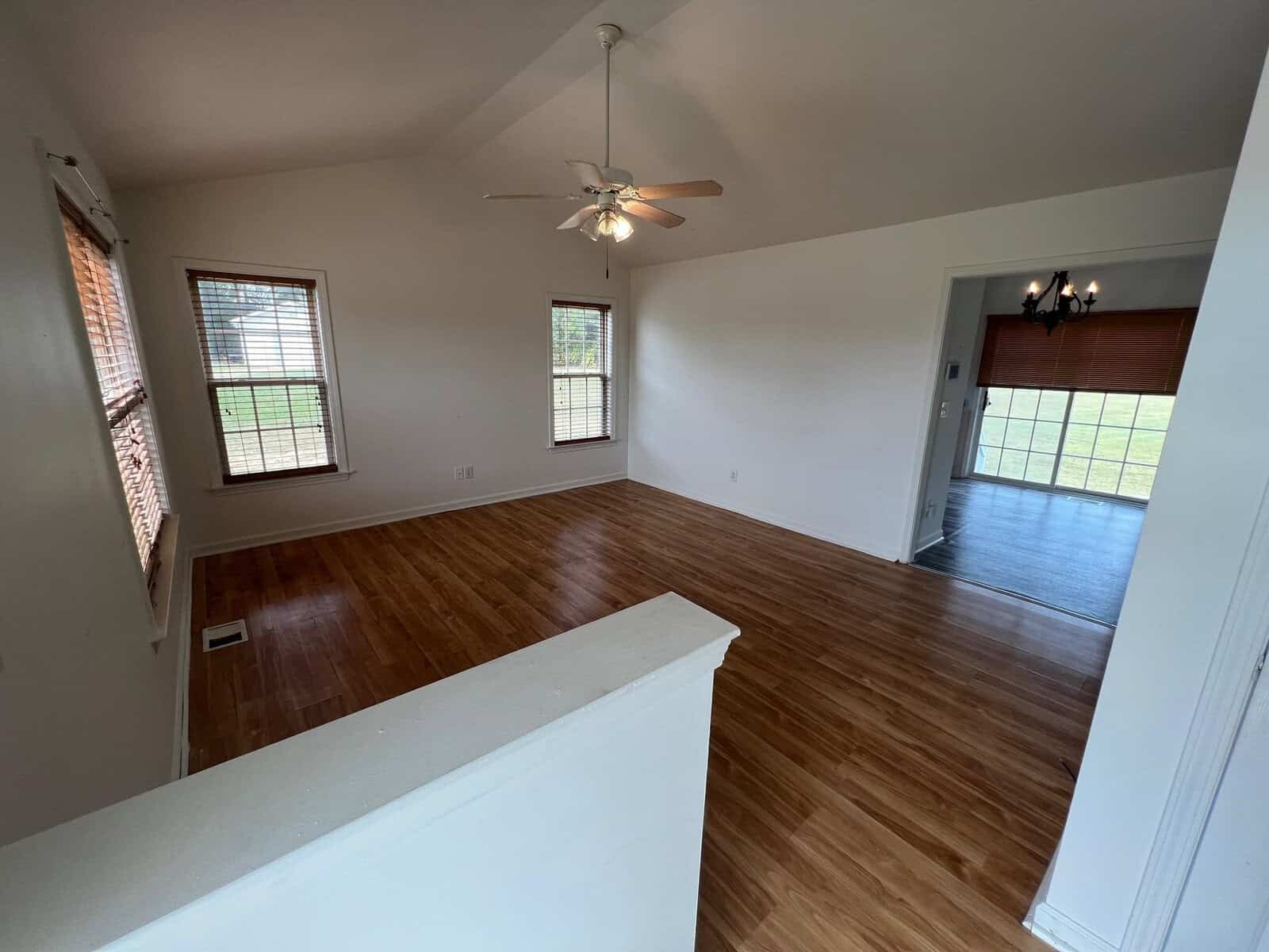 An empty room featuring hardwood floors, white walls, a ceiling fan, and multiple windows allowing natural light in, viewed from an elevated angle.