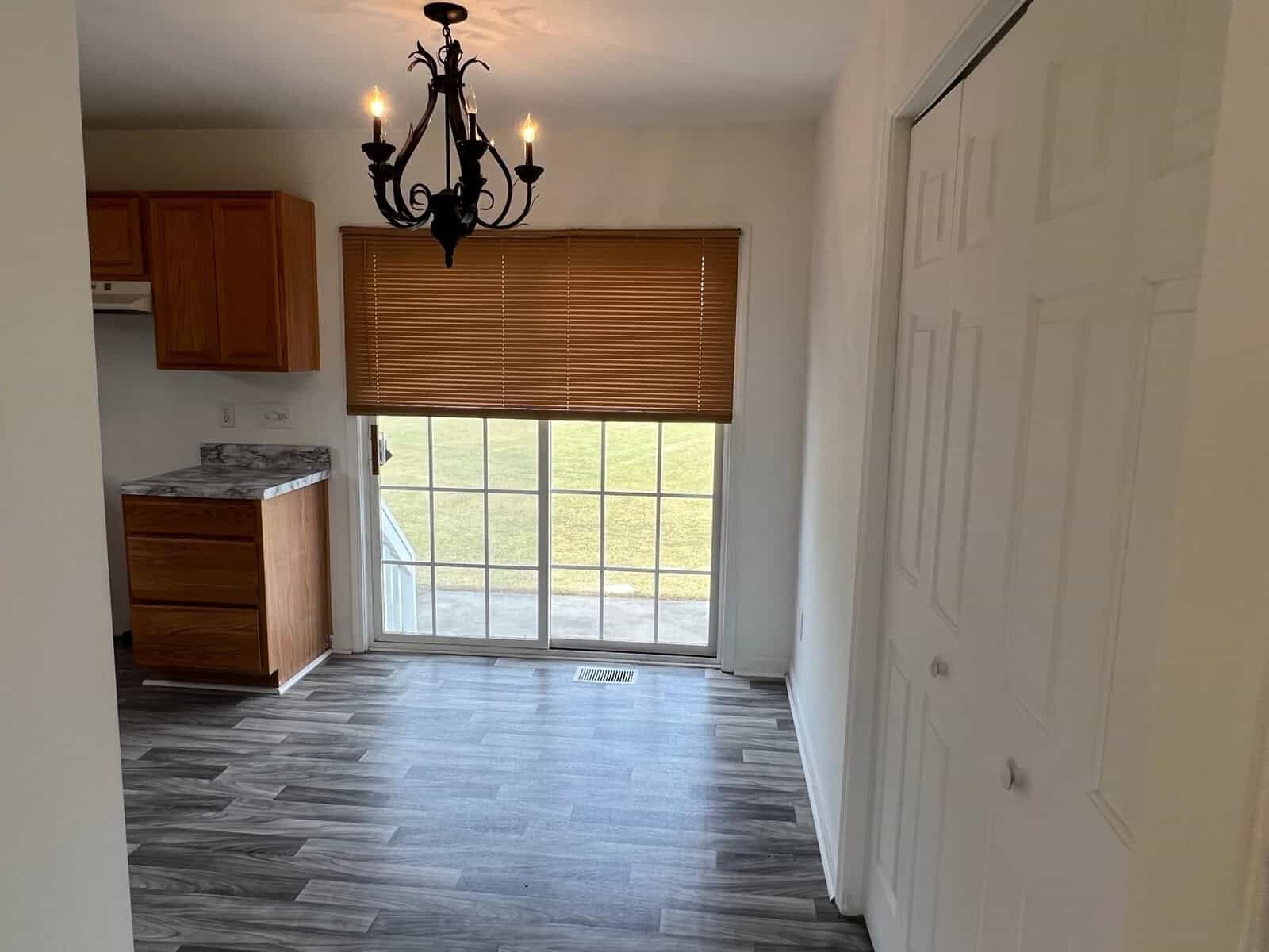 An empty room featuring a sliding glass door with a blind, a wooden kitchen cabinet to the left, a chandelier above, and a closed white door to the right, all above a grey laminate floor.