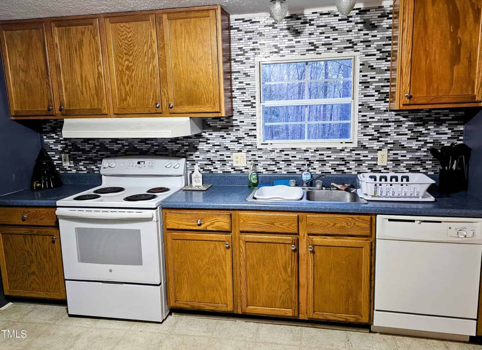 A kitchen with wooden cabinets and a blue countertop. Features include a white electric stove, white dishwasher, stainless steel sink with a drying rack, and a window above. The backsplash is a gray mosaic tile design.