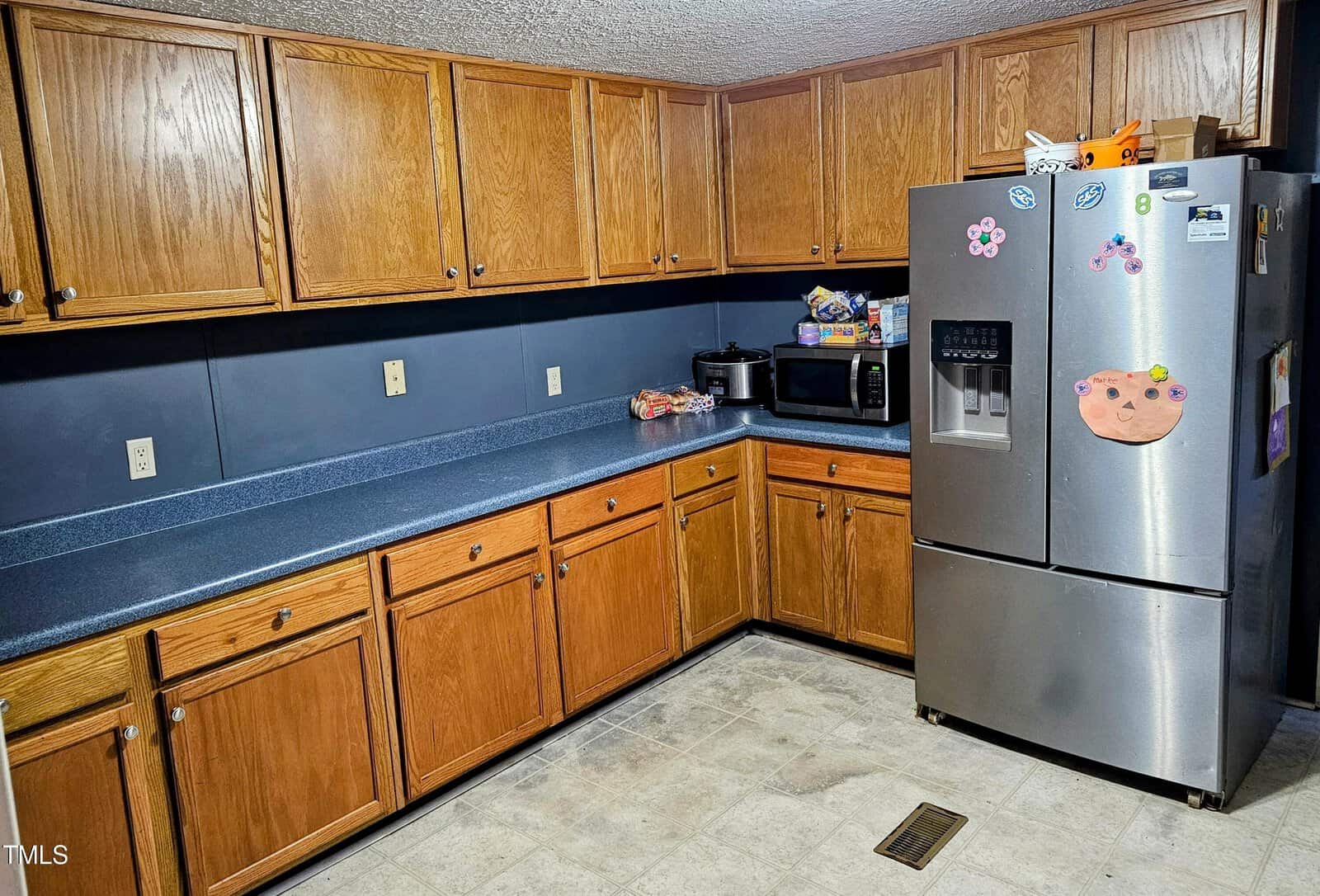 A kitchen with wooden cabinets, a blue countertop, and stainless steel refrigerator. The fridge has magnets and a drawing. A microwave and snacks are on the counter. The floor is tiled.