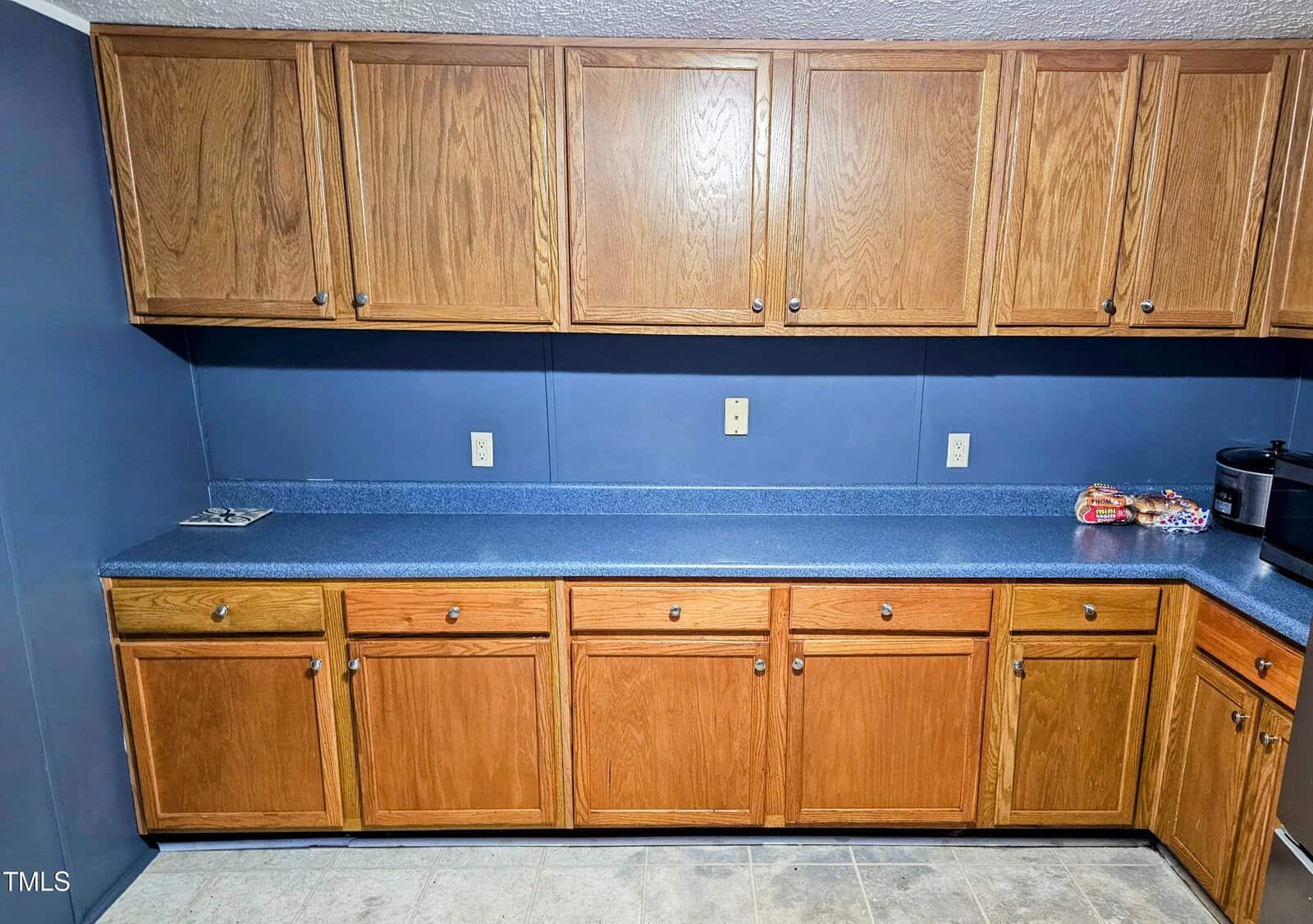 A kitchen with wooden cabinets and blue countertops. Upper and lower cabinets are closed. The blue backsplash matches the counter. Small kitchen appliances and snacks are on the counter to the right. The floor is light-colored tile.