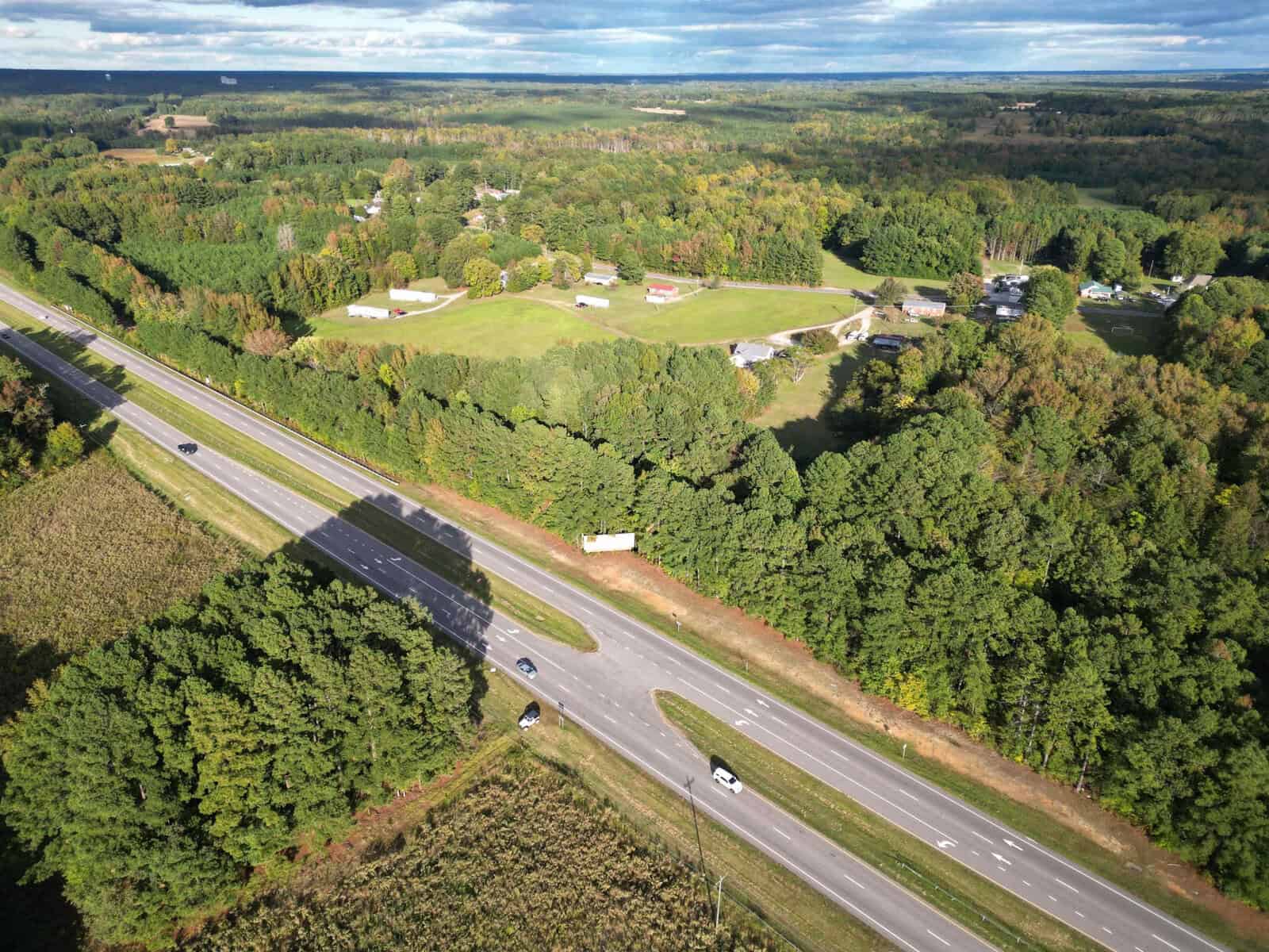 Aerial view of the 50 US 1 Highway winding through dense green forests near Kittrell, with scattered houses and fields. The road is lightly trafficked with a few cars visible. The landscape spreads towards a horizon of rolling hills under a partly cloudy sky.