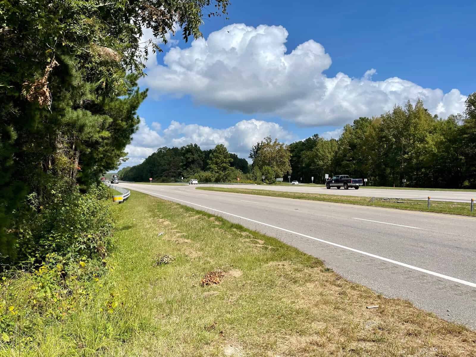 A rural highway lined with trees and greenery on a sunny day. The sky is filled with fluffy white clouds. Vehicles, including a truck and a car, are traveling on the road. The edge of the highway features grass and a metal guardrail.