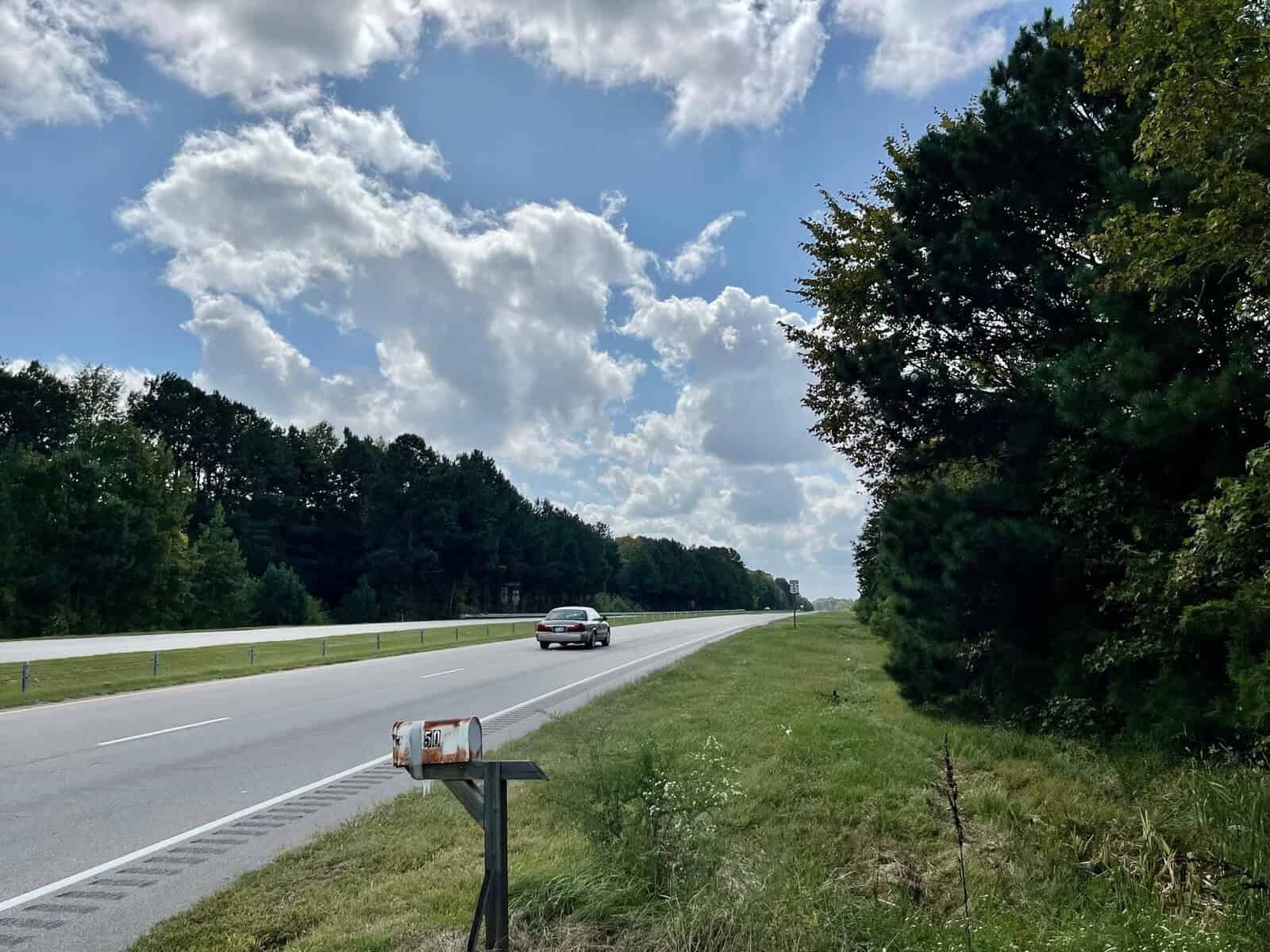 A rural road under a partly cloudy sky with a car driving away in the distance. The road is lined with trees on both sides and a rusty mailbox is visible in the foreground on the right side of the road.