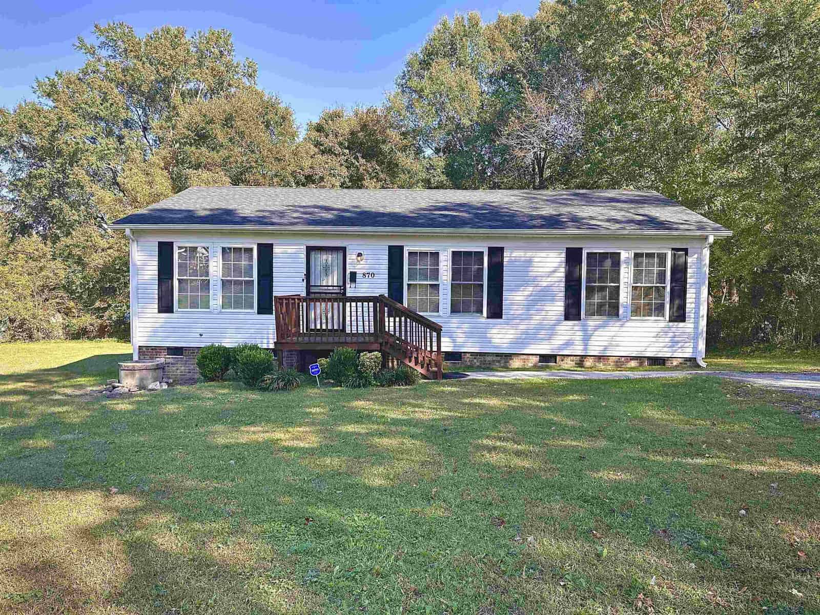 A white single-story house at 870 Bridgers Street, Henderson NC, with a gabled roof, featuring a small front porch with a wooden railing, surrounded by a lawn with trees in the background