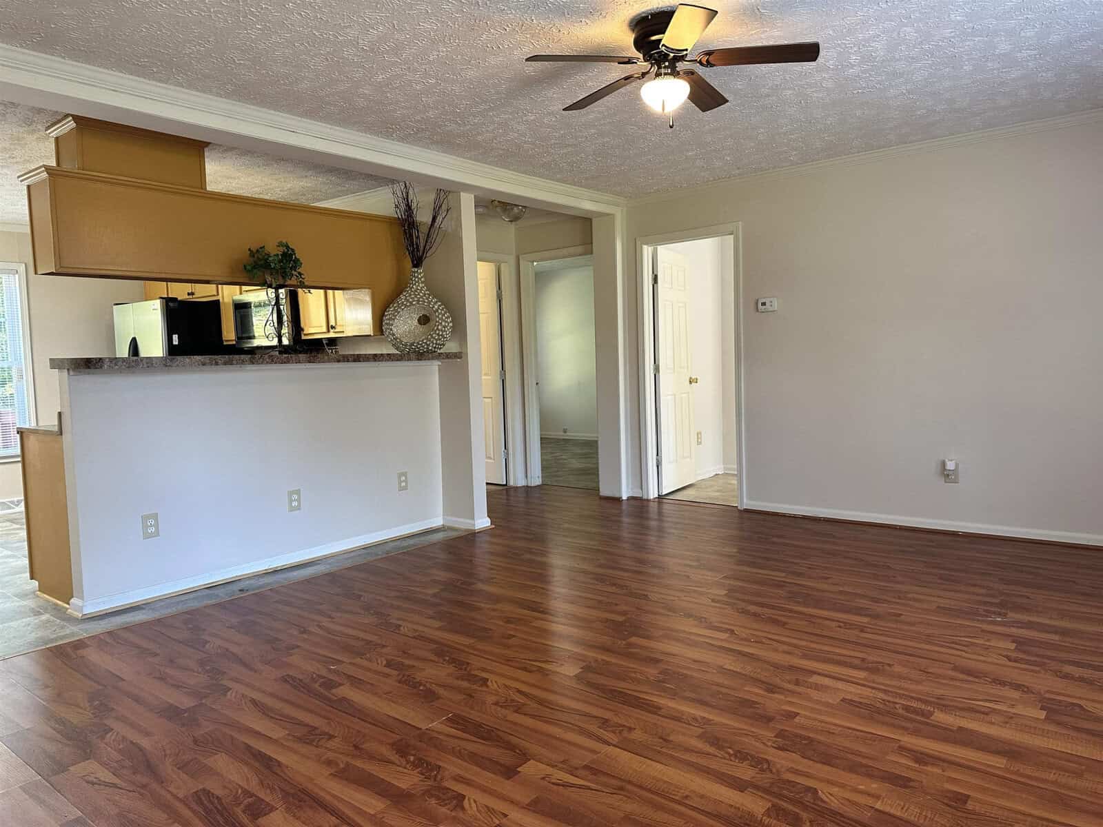 An empty room with hardwood flooring, featuring a kitchen counter with cabinets above it, a ceiling fan, and doors leading to other rooms.