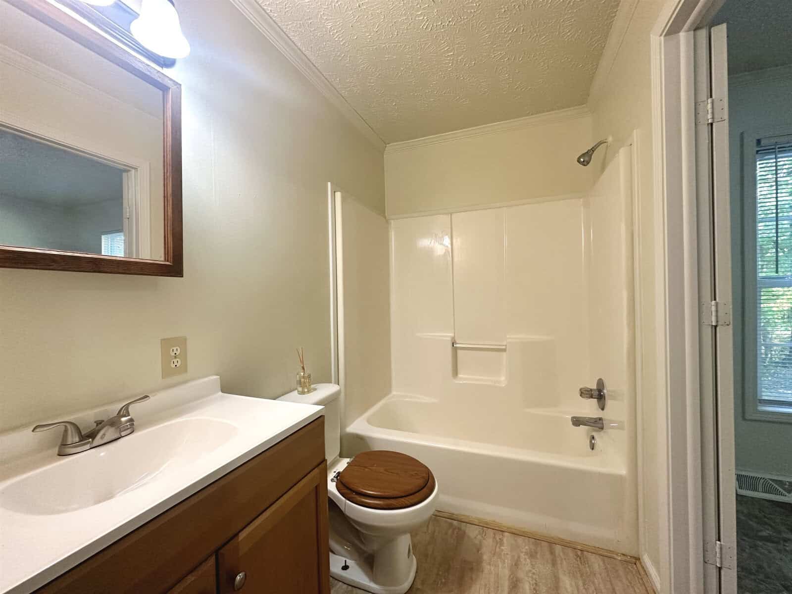 A small bathroom with a single vanity, mirror, toilet, and a shower-bathtub combo, featuring a window providing natural light.
