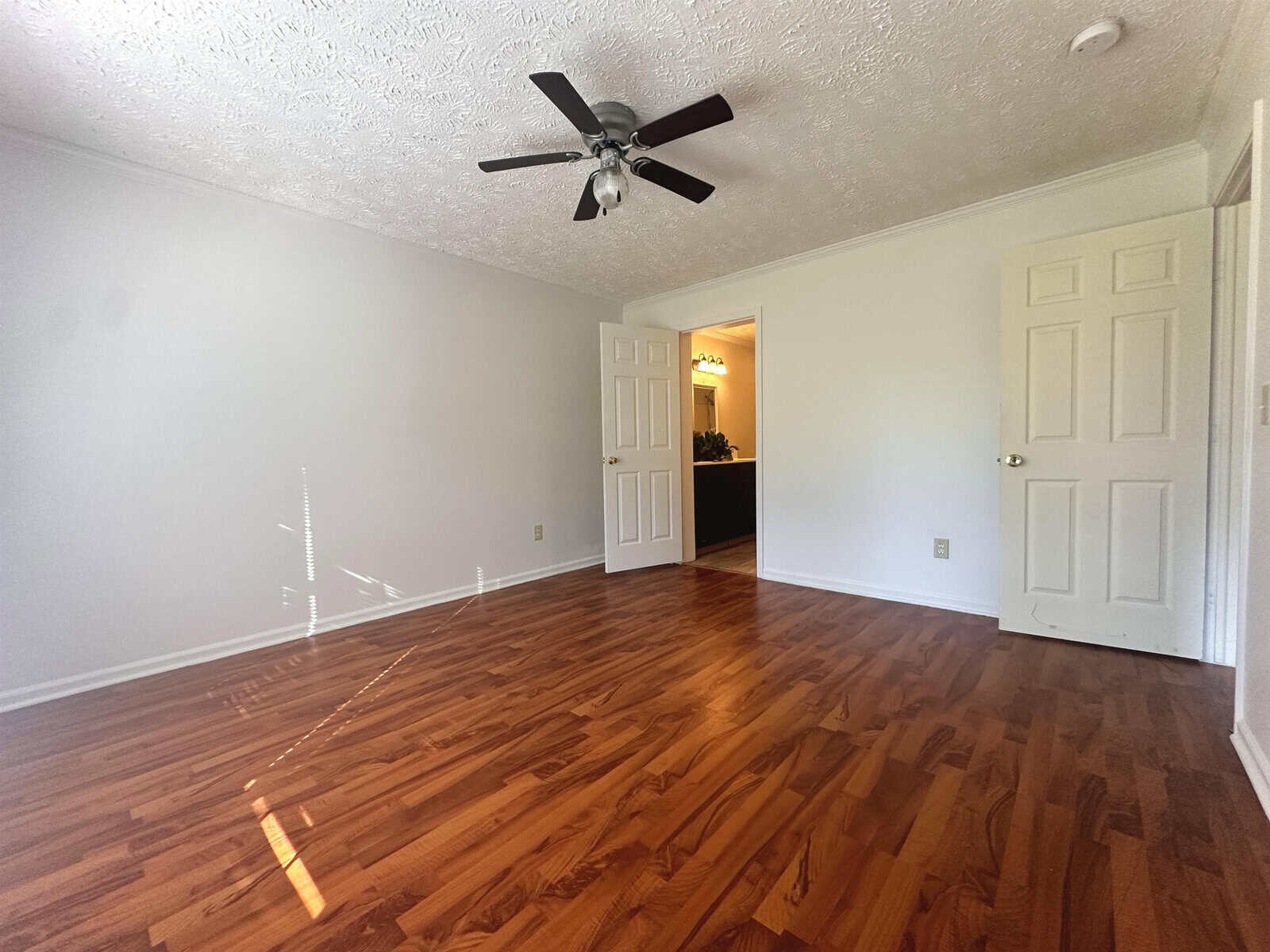 An empty room with polished wooden flooring, white walls, a closed white door to the right, and an open doorway leading to another room, under a ceiling fan with lights off.