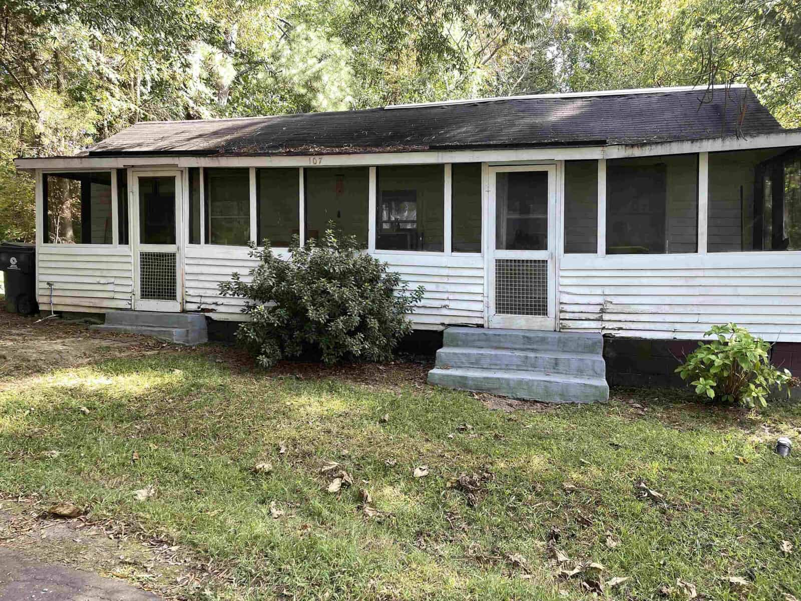 Nestled at 107 5th Street in Oxford, this small weathered white house features a screened-in porch and a shingled roof. Surrounded by green trees and grass, two concrete steps lead to the porch, with bushes adorning the front.