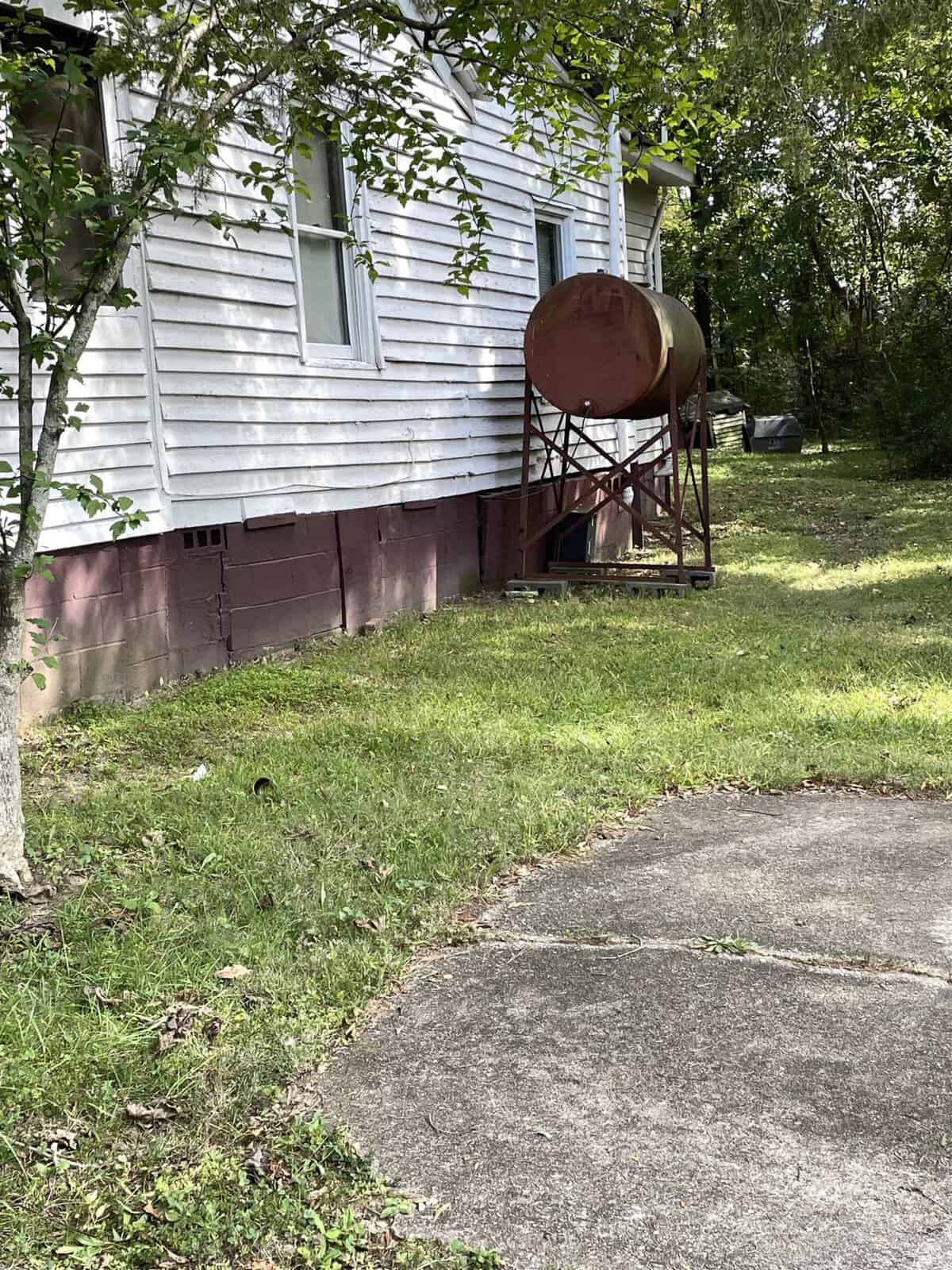 A white house with a brick foundation has an old, rusted oil tank on a metal stand next to it. The yard has green grass and a concrete path in the foreground, surrounded by trees and bushes.