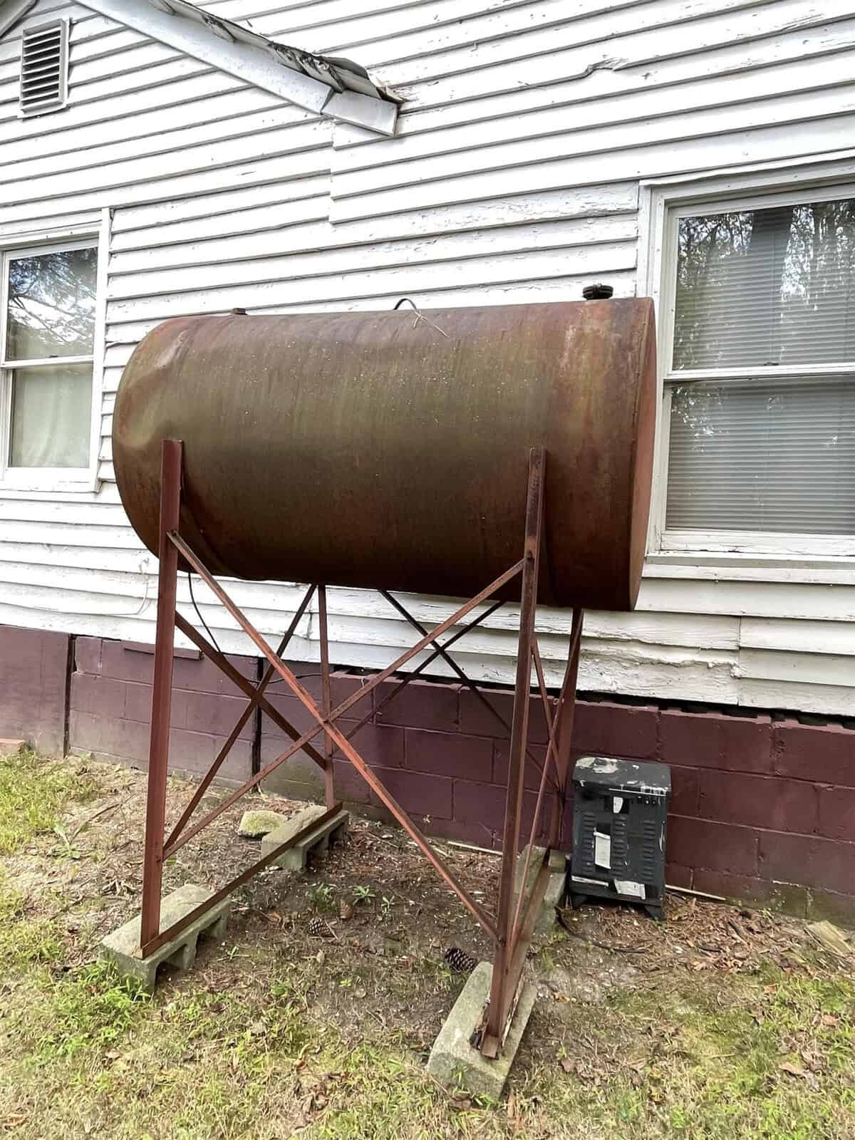 A large, rusty oil tank is elevated on a metal stand next to a white wooden house. A window is visible on the house's side, and there is a black object, possibly a container, on the ground next to the tank.