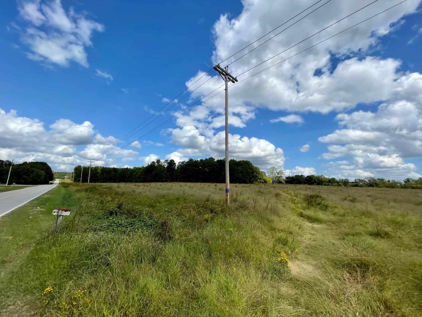 A rural landscape featuring a lush grassy field with a utility pole and wires, under a vibrant blue sky with fluffy white clouds. a road runs along the left side, with a clear view and no vehicles.