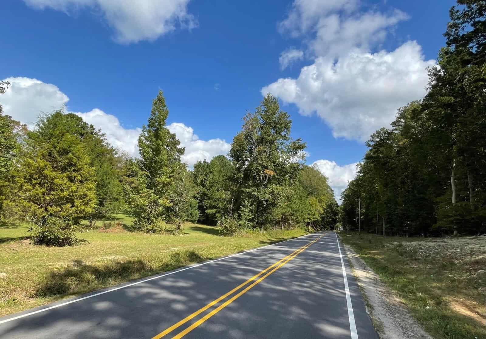 A rural road with double yellow lines stretches into the distance, bordered by grassy fields and lush green trees under a bright blue sky with scattered, fluffy white clouds.