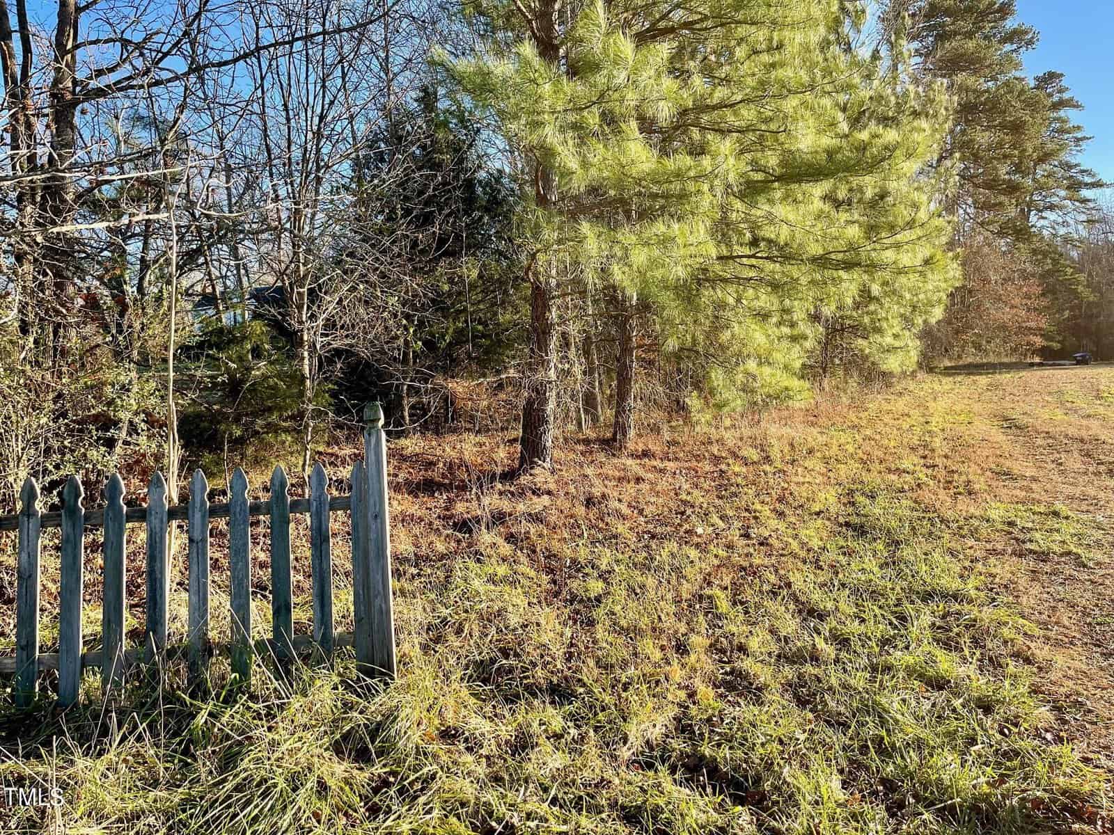 A rural scene shows a partially broken, weathered wooden fence in the foreground along 50' Kittrell Road, with dry grass and various trees including conifers in the background. The sun casts long shadows, indicating early morning or late afternoon.