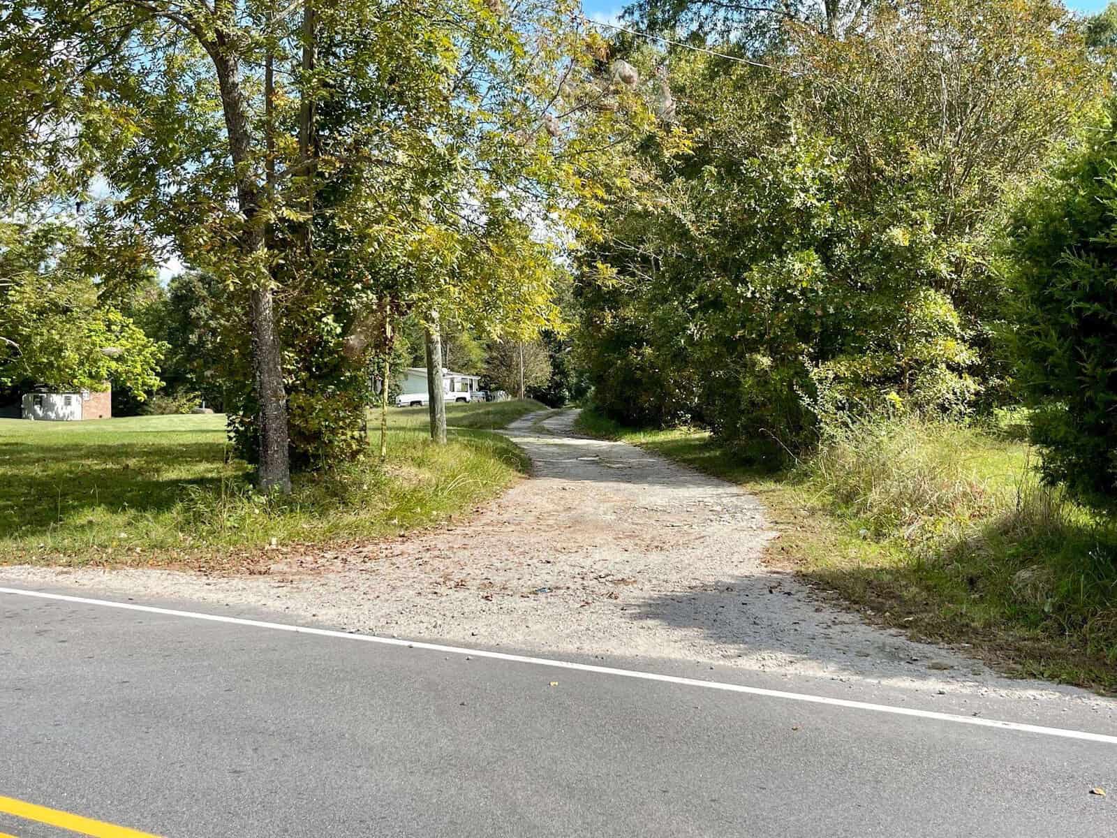 A narrow gravel driveway leads from a paved road into a wooded area with lush green trees and foliage. In the distance, a white house at 011 E Chavis Lane in Kittrell is partially visible, surrounded by greenery on a clear day.