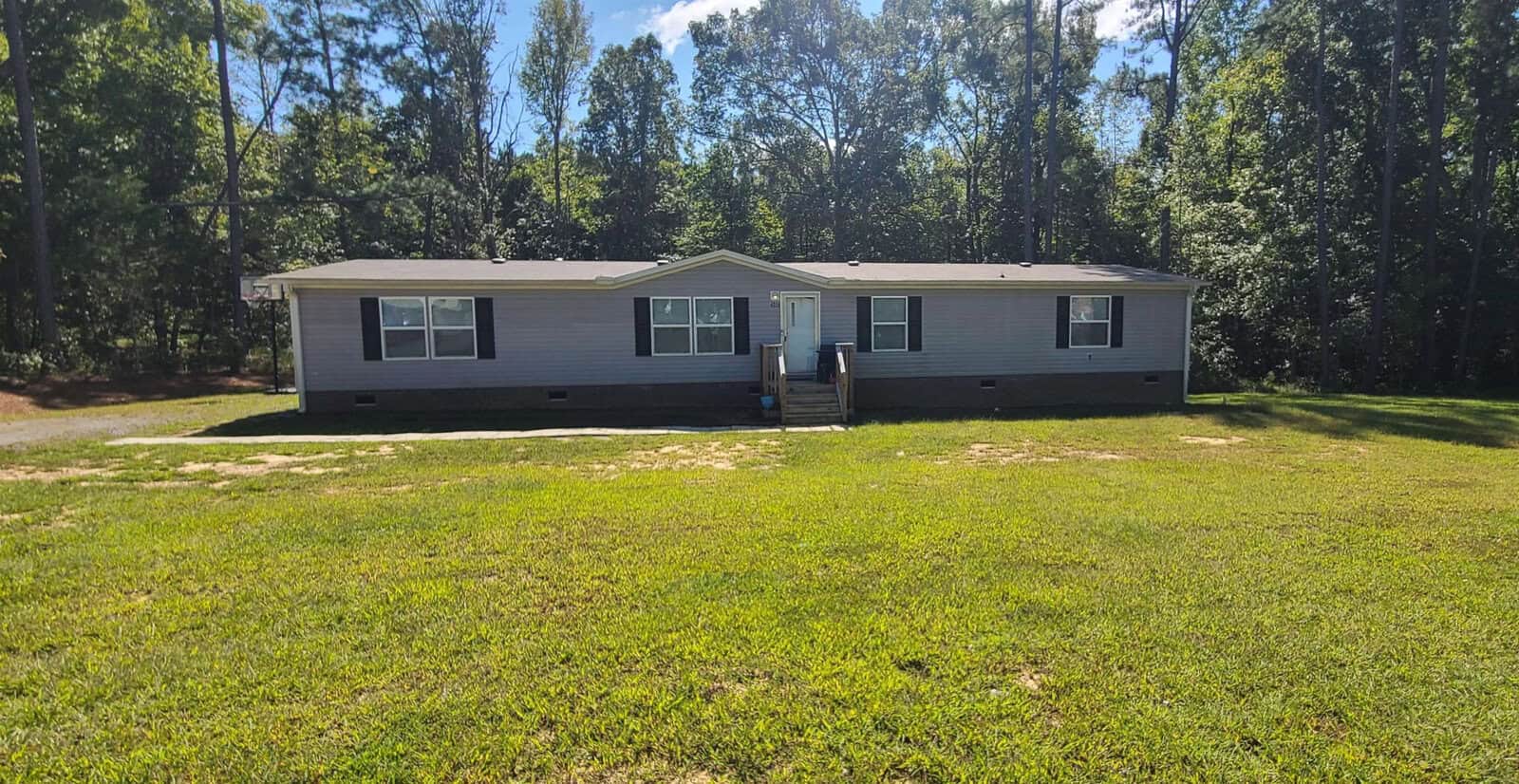 Nestled at 4056 Shannon Drive, Oxford, this single-story gray house features a white front door and multiple windows. It stands gracefully amid lush green grass and tall trees under a clear blue sky. Steps elegantly lead to the entrance at the center of the building.