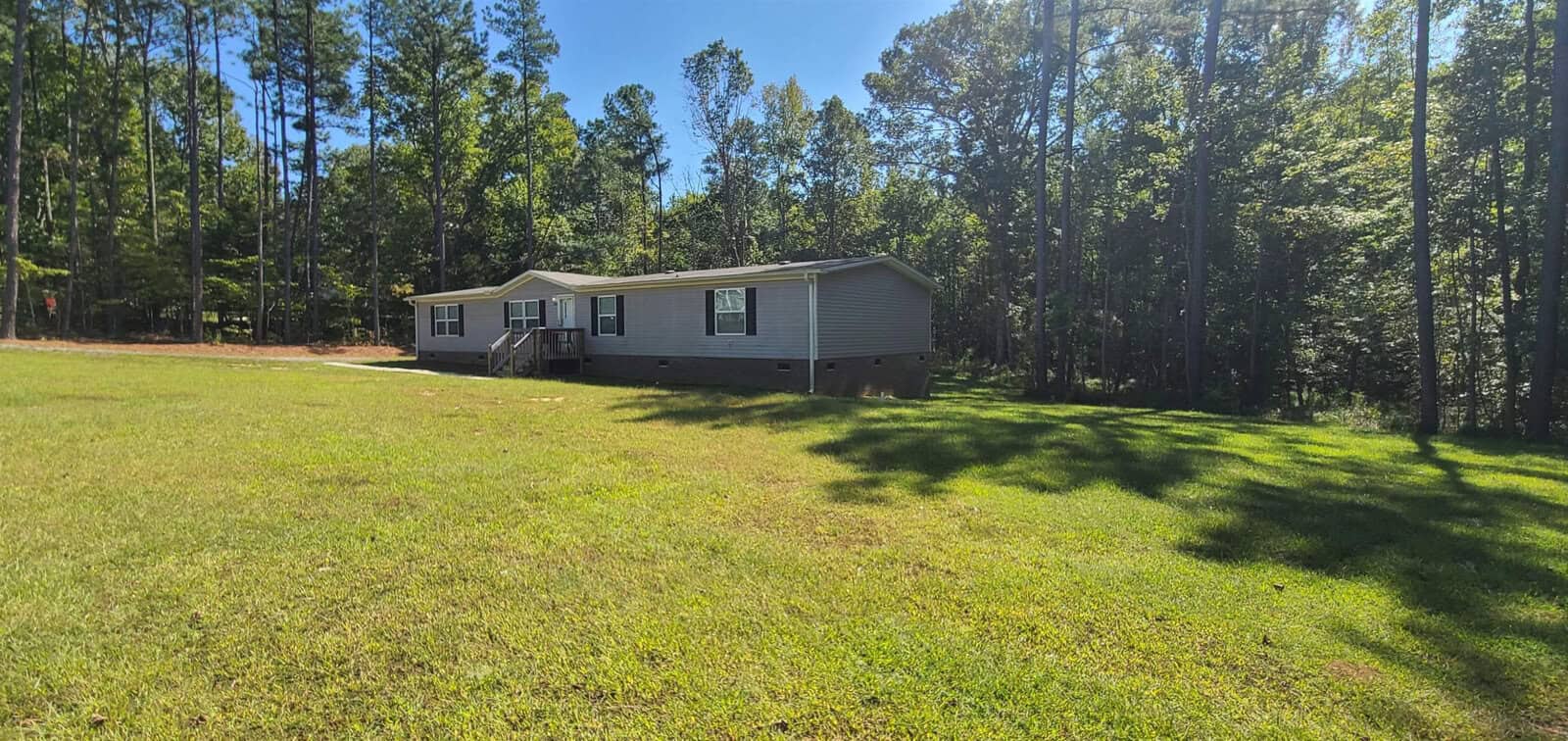A gray mobile home sits on a spacious lawn surrounded by tall trees under a clear blue sky. Sunlight casts soft shadows across the grass.