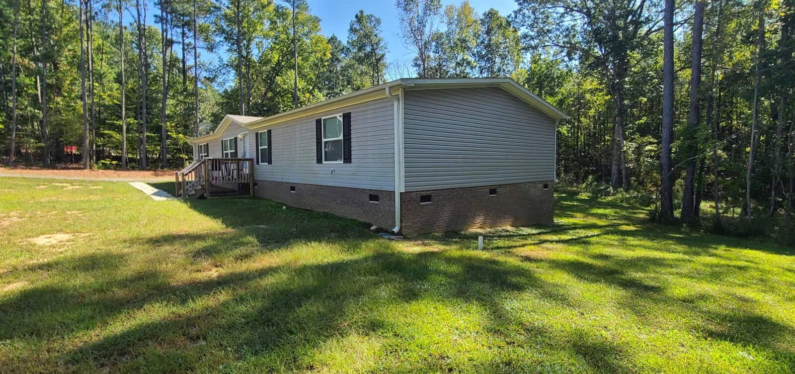 A single-story gray mobile home with a brick foundation is situated in a grassy yard surrounded by tall trees. The home has several rectangular windows and a small wooden porch at the front.