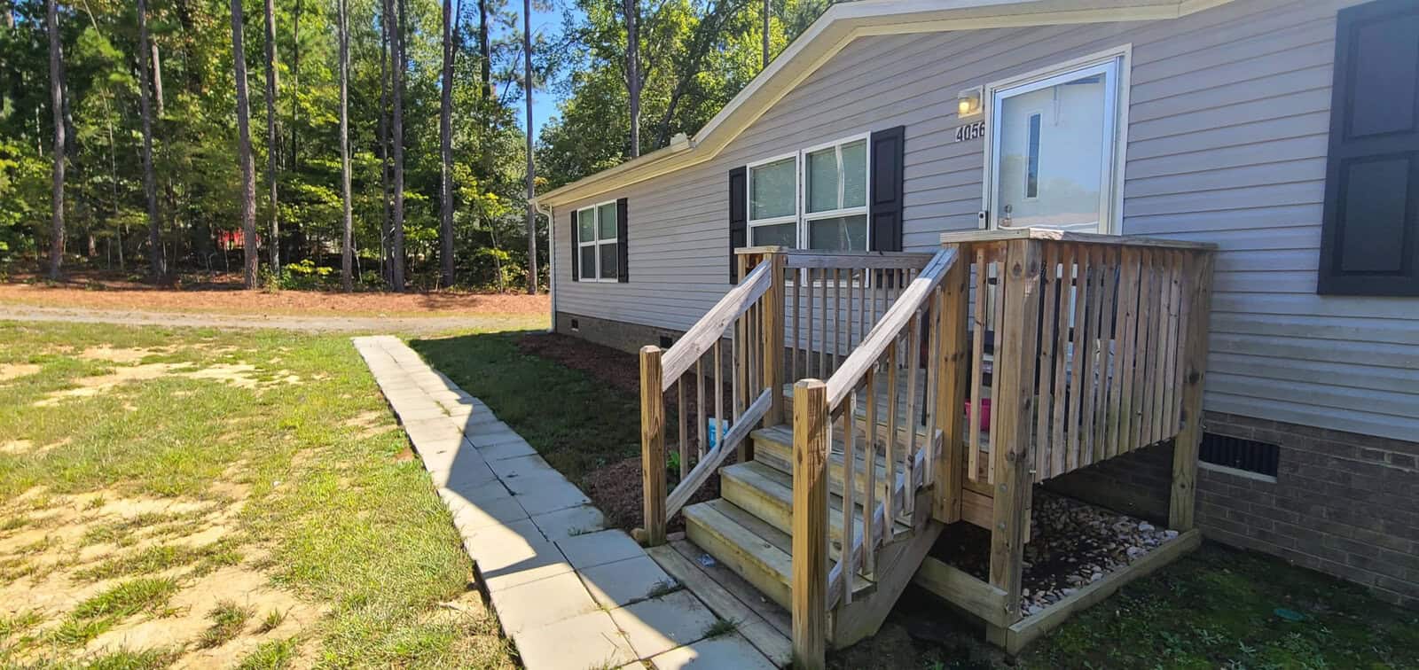 A small beige mobile home with a wooden porch and steps. There's a path leading to the entrance, surrounded by a grassy lawn and tall trees in the background. The scene is bright and sunny.
