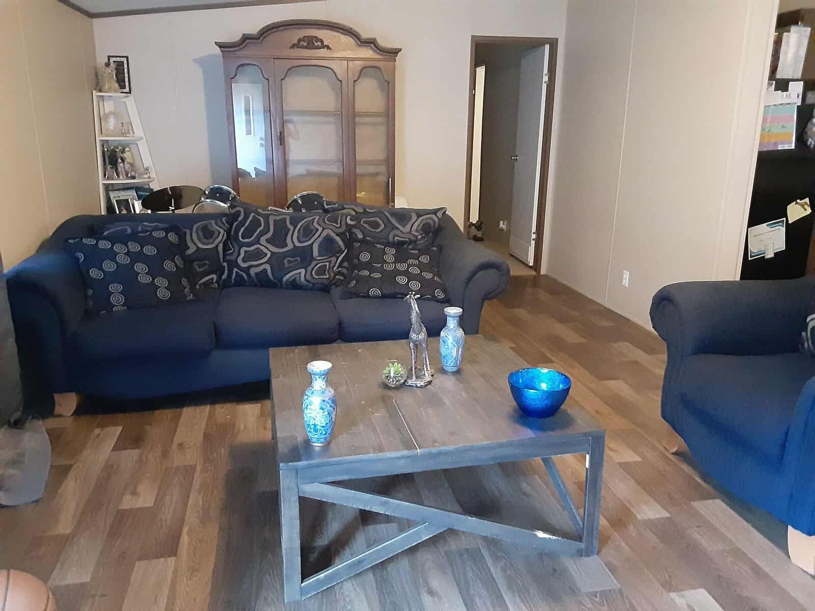 A cozy living room with a dark blue sofa and matching armchair, adorned with patterned cushions. A wooden coffee table displays decorative vases and a blue bowl. A large wooden cabinet is in the background. The floor is wood laminate.