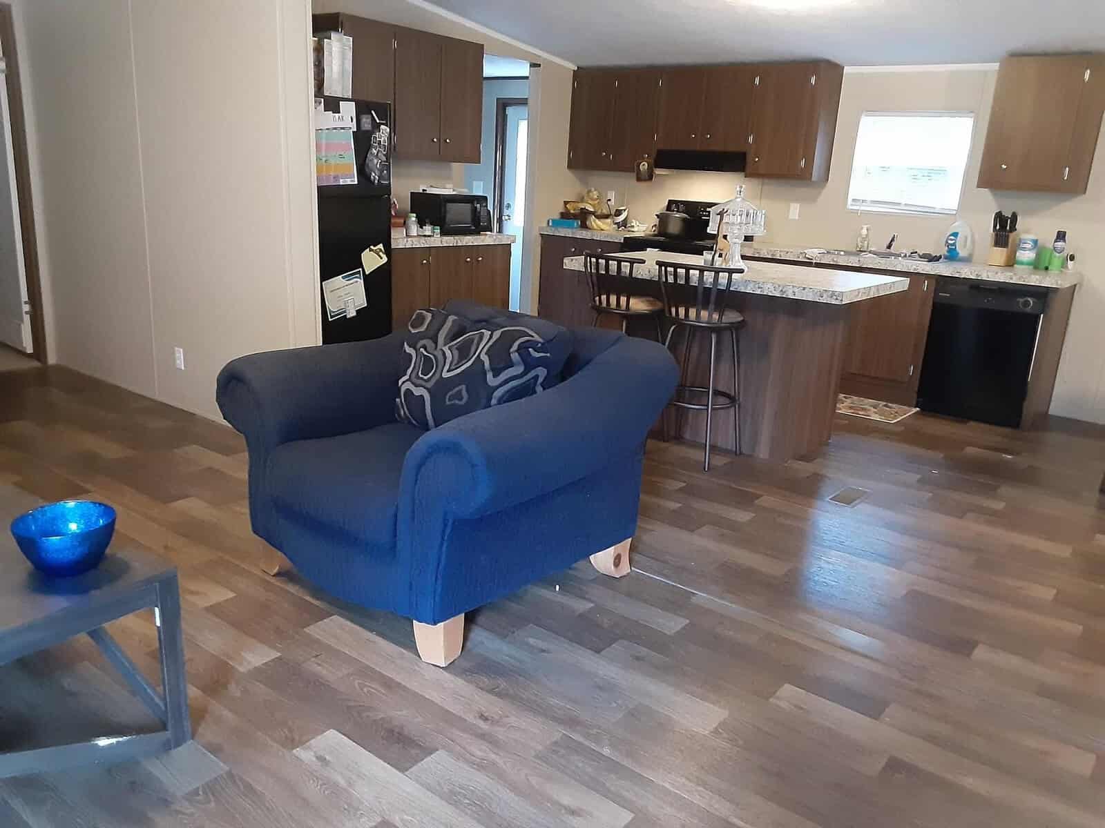 A cozy living room and kitchen with wooden floors. A blue cushioned armchair faces a small table with a blue bowl. Wooden kitchen cabinets and a black refrigerator are in the background, with assorted items on the countertops.