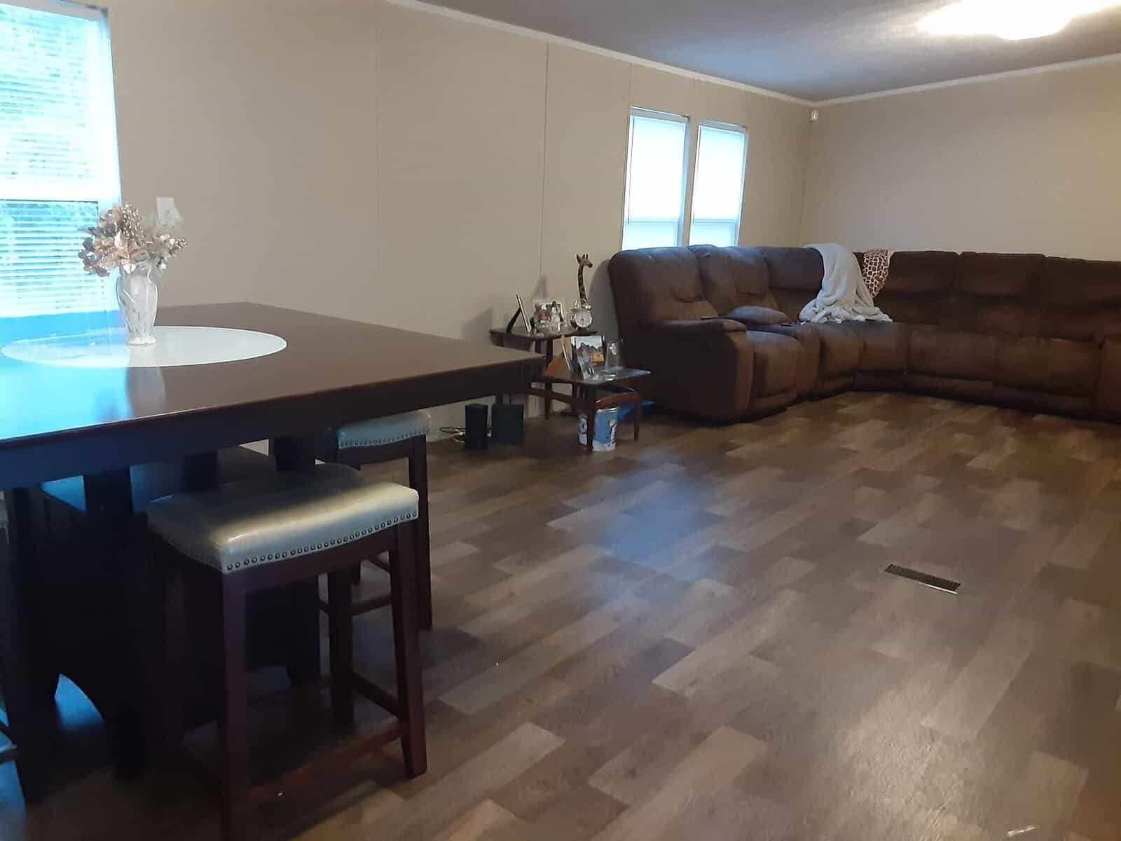 A spacious living room with wooden flooring, featuring a brown sectional sofa with a blanket draped over it. A high table with two stools is in the foreground, and a vase with flowers sits on the table. Natural light enters through the windows.