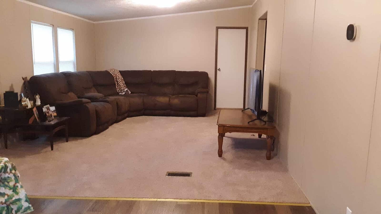 A spacious living room with beige walls and carpet, featuring a large brown sectional sofa. A flat-screen TV sits on a wooden coffee table. A small side table holds framed photos and a blanket is draped over the sofa.