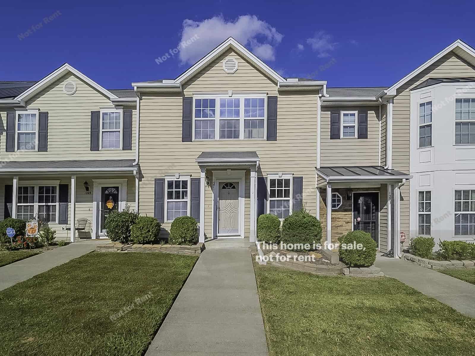 A row of beige and light yellow townhouses under a clear blue sky. The central townhouse at 1271 Quail Circle boasts a small porch with shrubs on either side. A sign in front reads, "This home is for sale, not for rent," right in the heart of Creedmoor.