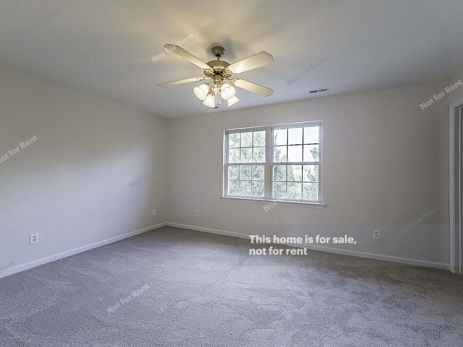 An empty, carpeted room with beige walls features a ceiling fan with lights and a large window letting in natural light. Text on the walls and floor states, "This home is for sale, not for rent.