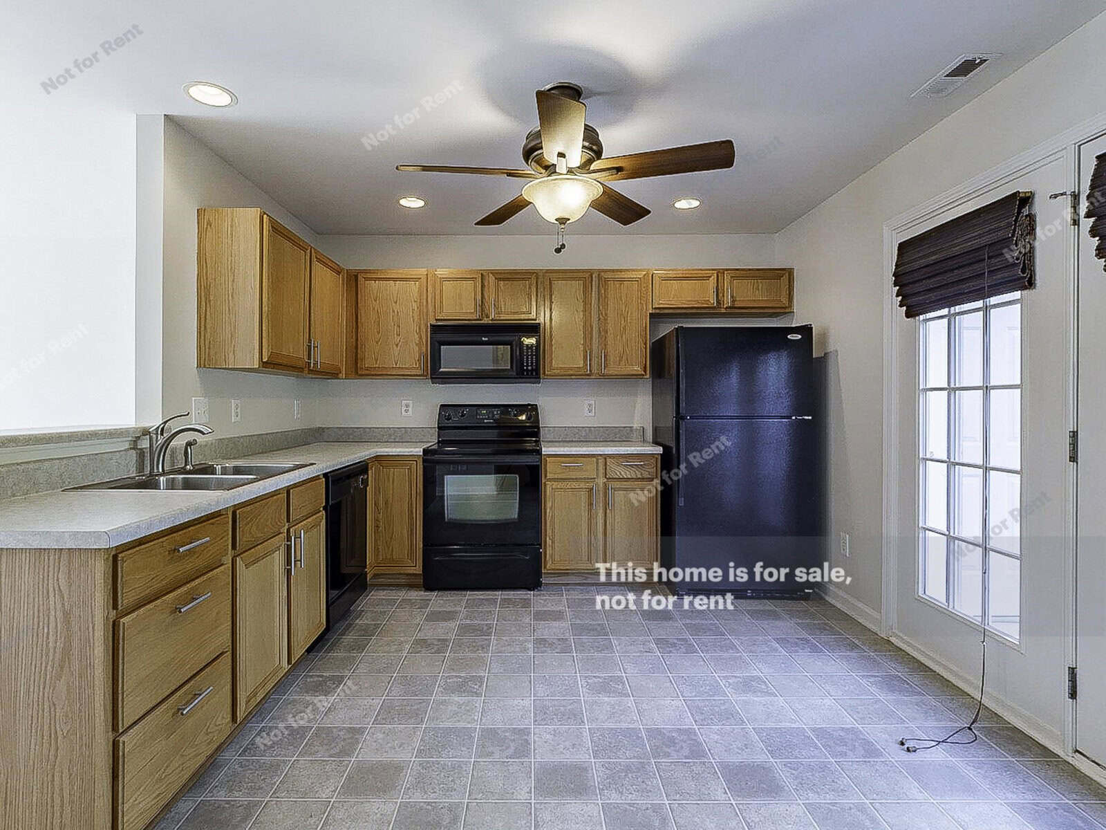 A modern kitchen with wooden cabinets and black appliances, including a refrigerator, oven, and microwave. A ceiling fan is centered above, and there's a windowed door on the right. The floor features gray tiles.