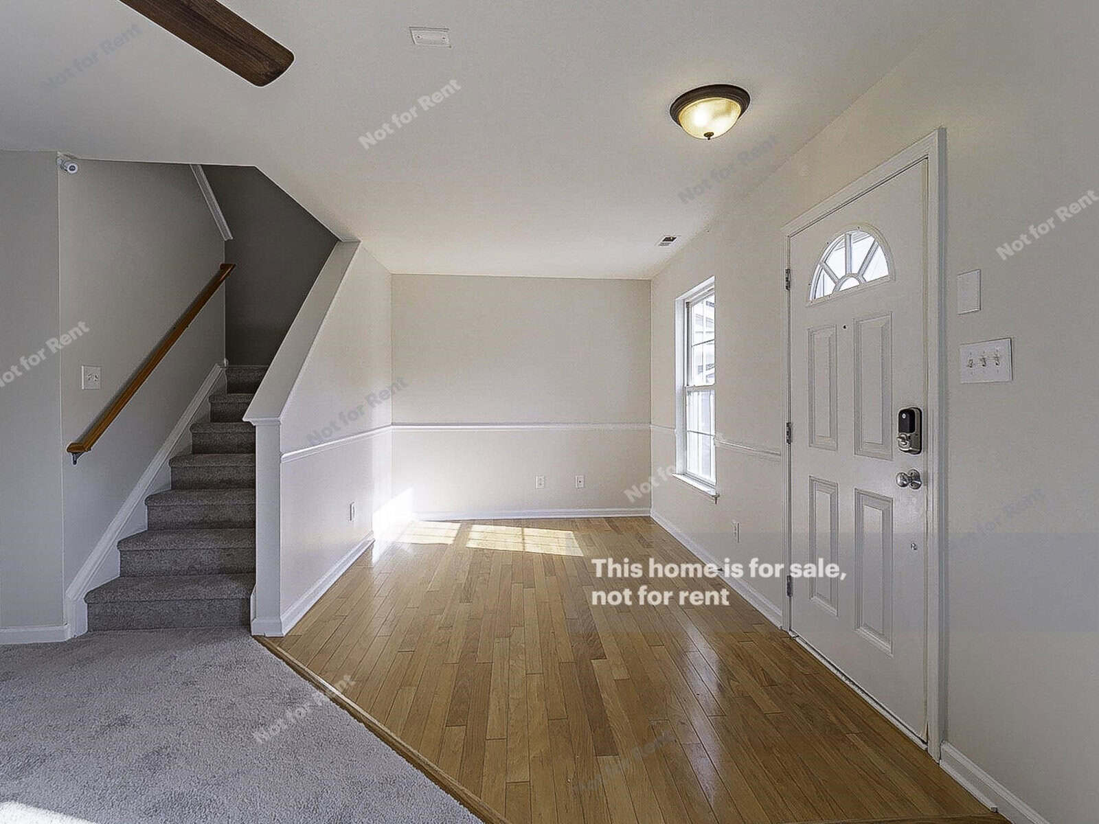 A bright room with wooden flooring and a carpeted staircase. Sunlight streams through a window near the white front door. Text on the image indicates that the home is for sale, not for rent.
