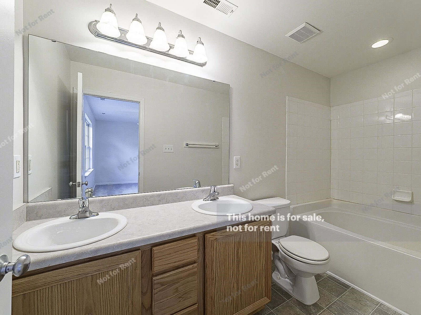 A bathroom with a large mirror above a dual-sink vanity, wooden cabinets, and a counter. The room features a bathtub with white tile backsplash, a toilet, and overhead lighting. A door leads to an adjacent room. Text reads "This home is for sale, not for rent.
