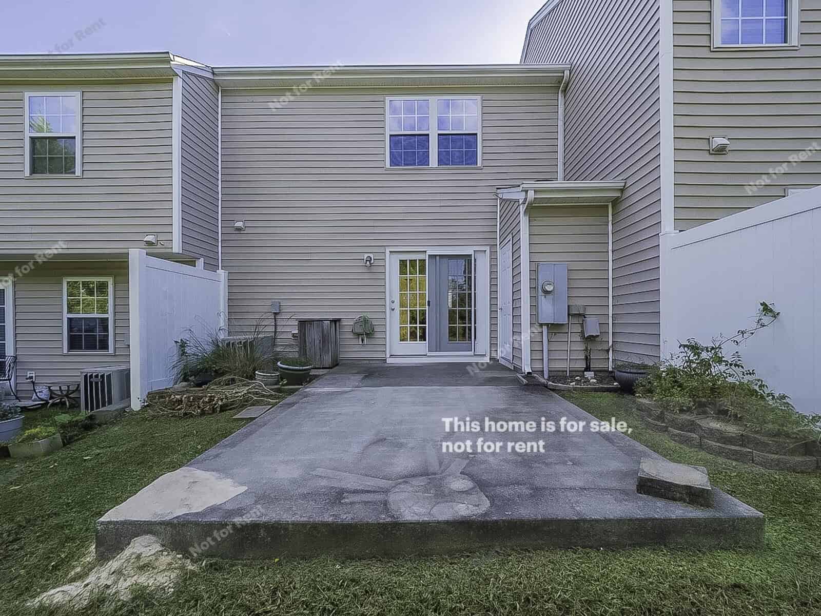 A two-story house exterior with beige siding. The backyard features a concrete patio, small garden, and grass. There are sliding glass doors leading inside. Text on the image reads, "This home is for sale, not for rent.