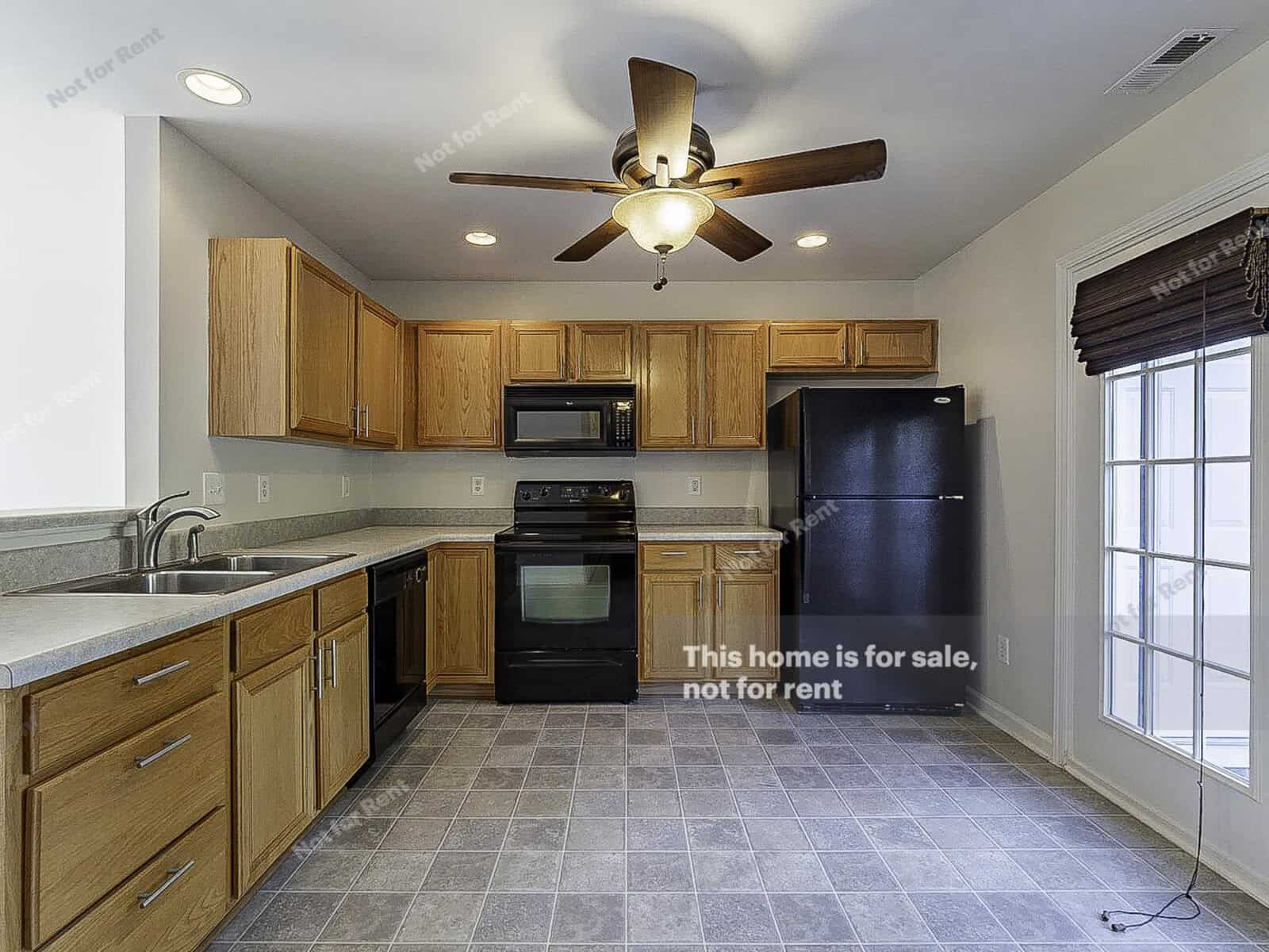 A modern kitchen with wooden cabinets, black appliances, and a ceiling fan. The room features a tiled floor and a window with blinds. Text on the image states, "This home is for sale, not for rent.