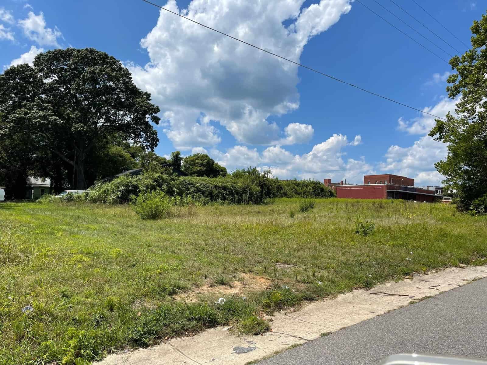 A grassy vacant lot bordered by trees and shrubs under a partly cloudy sky with buildings in the background.