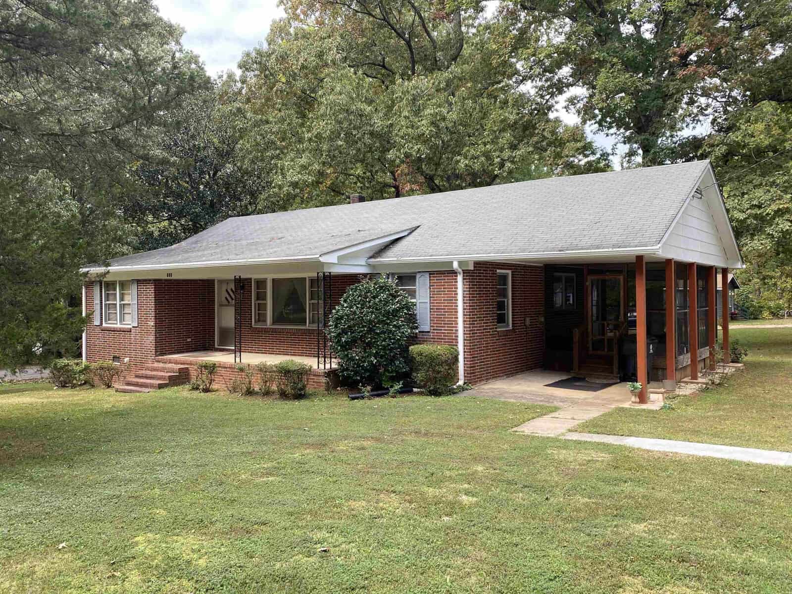 A single-story red brick house with white trim, a gray shingled roof, and a covered front porch surrounded by green lawn and trees.