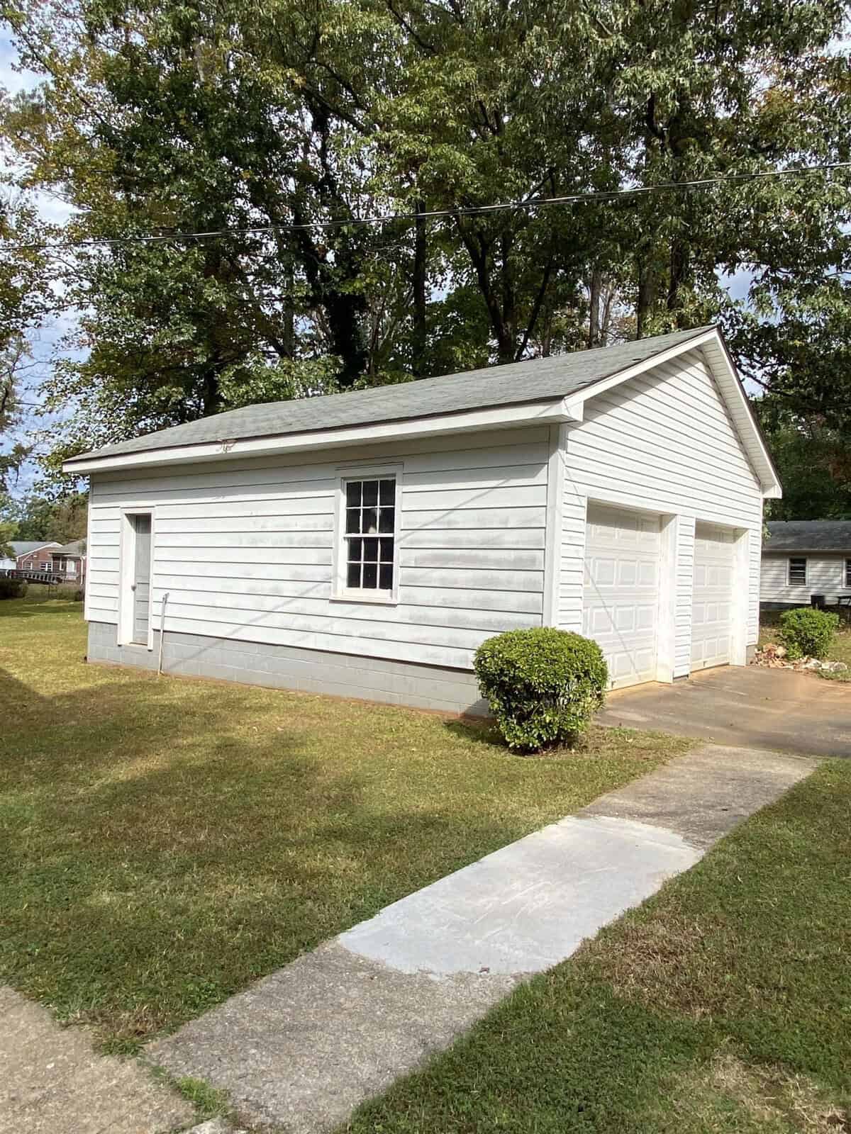 A small, white, single-story garage building with a single window and a single garage door, situated beside a concrete pathway and surrounded by grass and a trimmed bush, under a clear sky.
