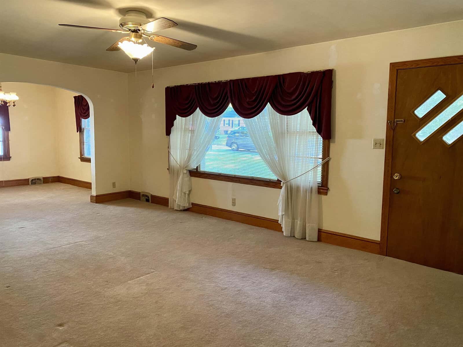 A well-lit, empty room featuring a large window with sheer curtains and red drapes, a ceiling fan, and beige carpeting. a wooden door with small glass panels is visible on the right.