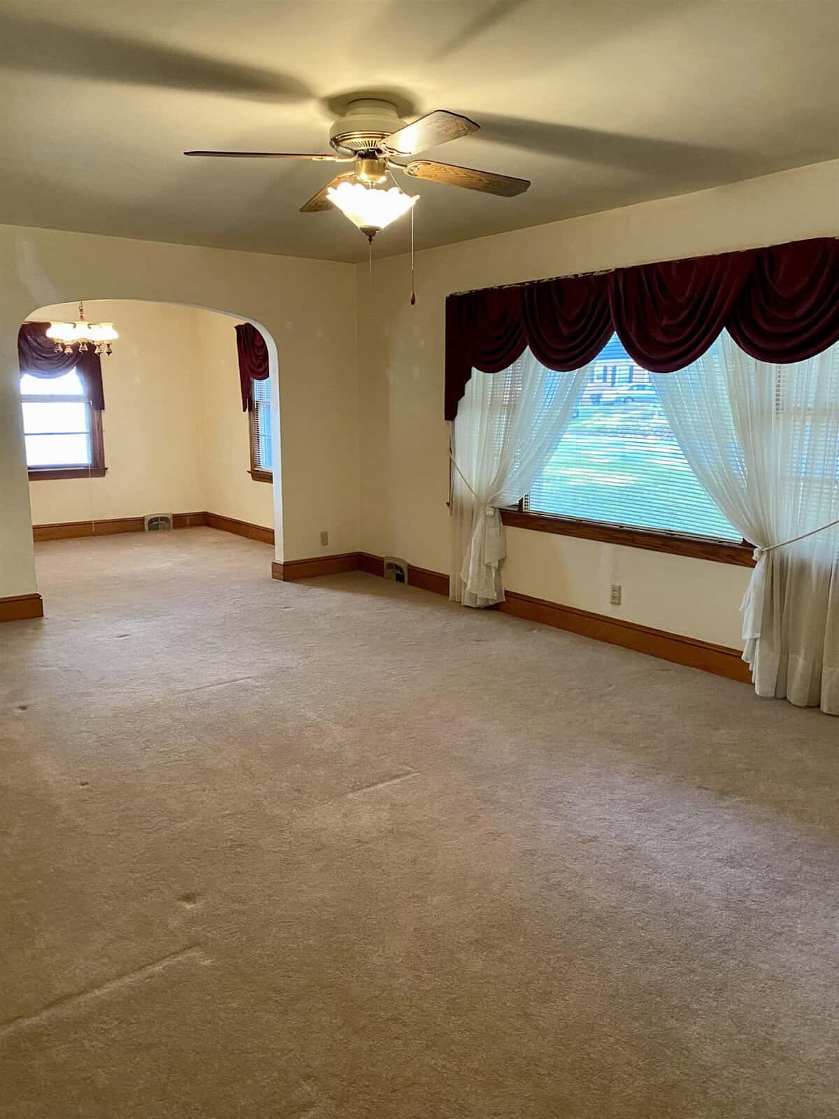 An empty room with beige carpeting, a ceiling fan, and windows dressed with red valances and sheer white curtains.