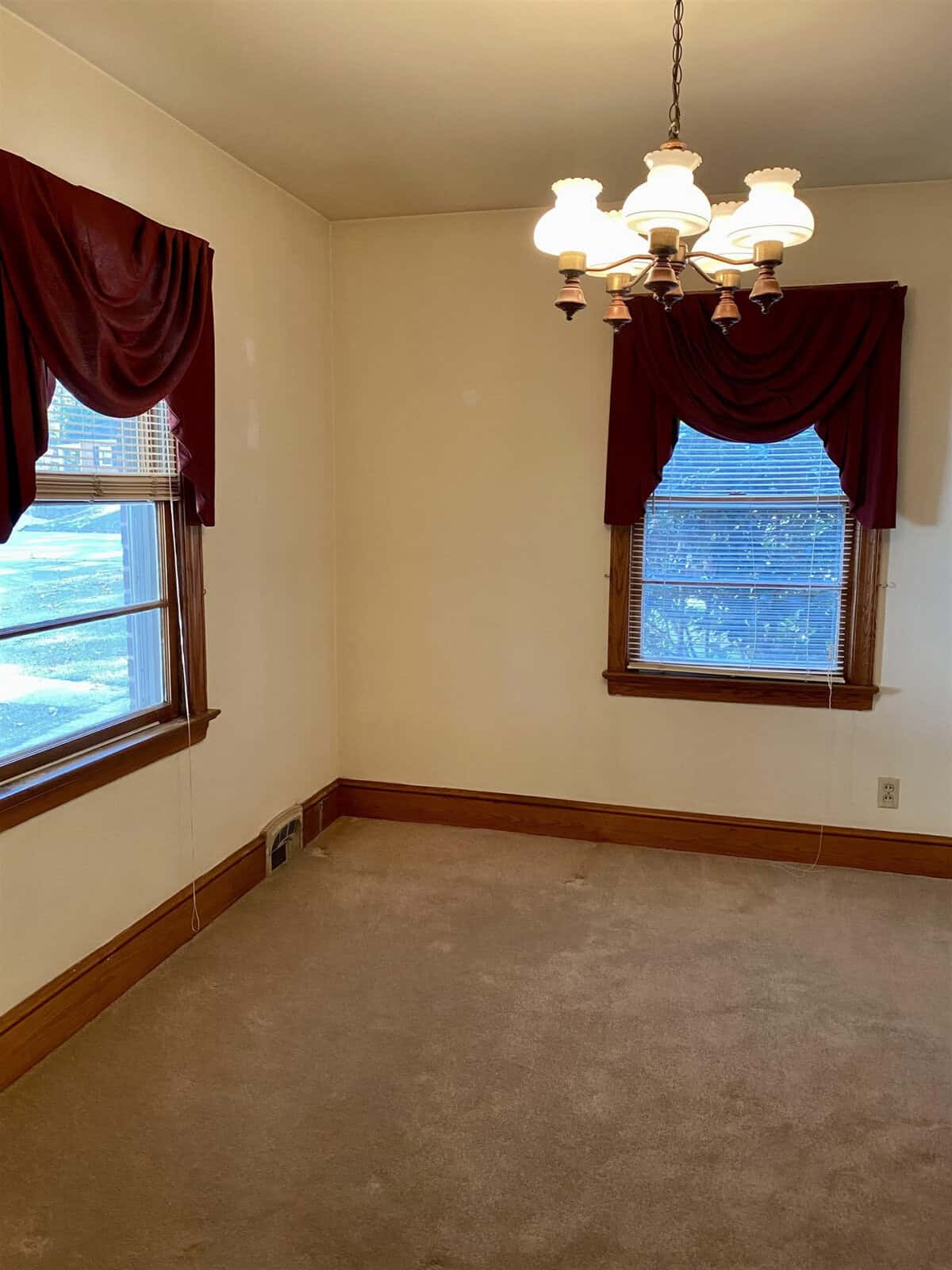 An empty room with beige walls and brown carpeting, featuring two windows with red drapery and a chandelier.