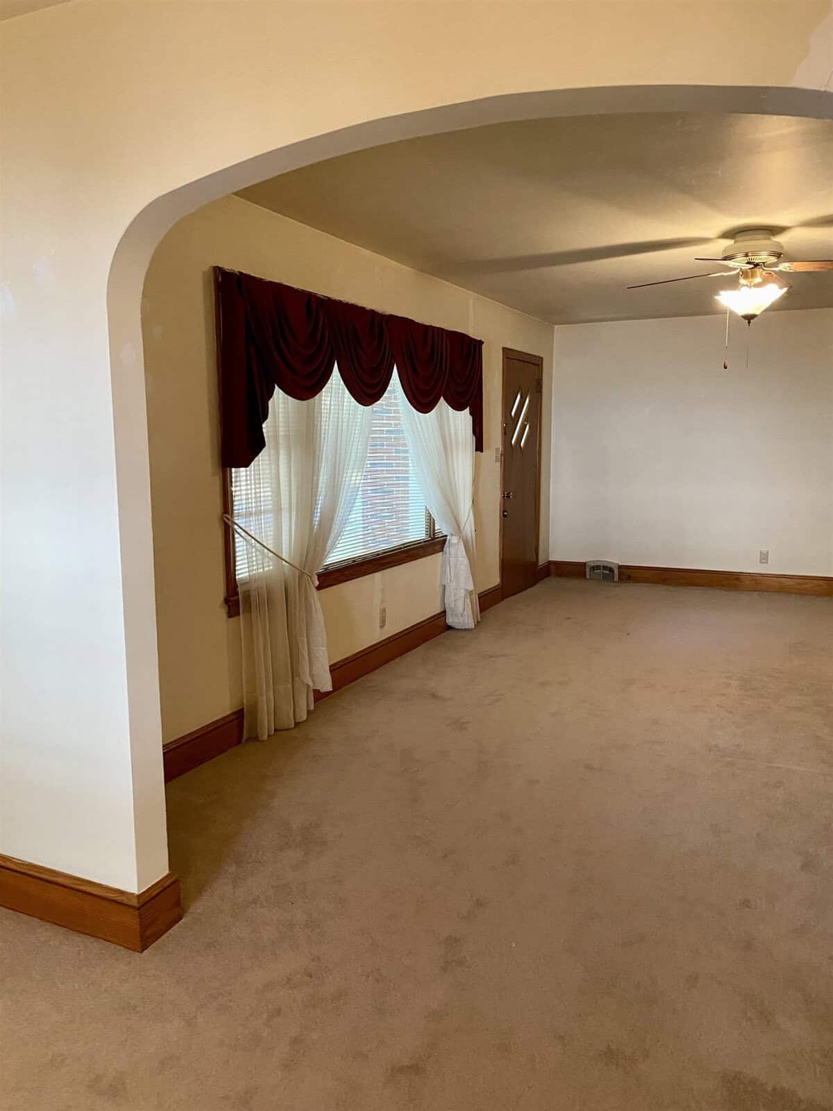 Empty room with a tan carpet, white walls, arched doorway, ceiling fan, and a bay window draped with maroon valances and white sheer curtains.