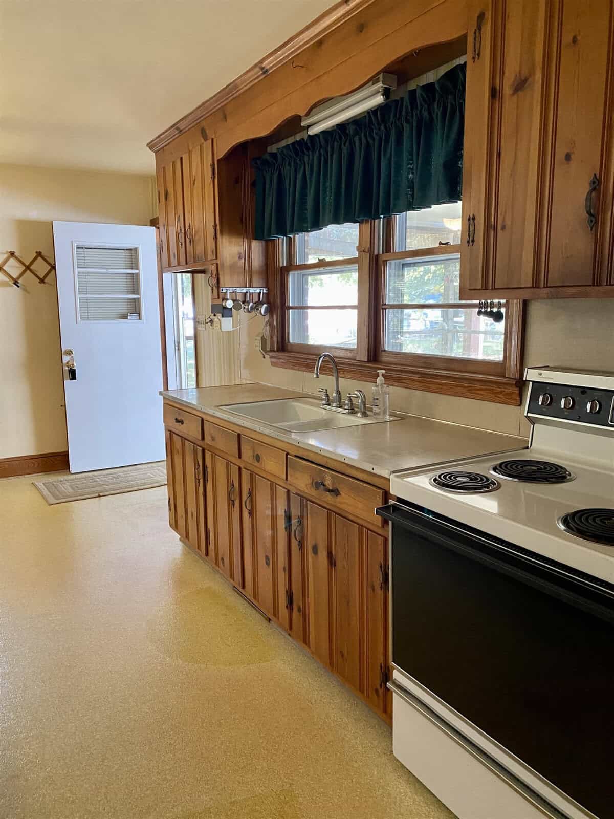 A traditional kitchen interior with wooden cabinets, a white double sink below a window with green curtains, and a white stove to the right.