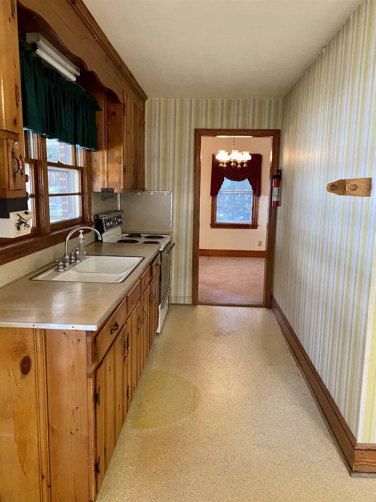 Narrow kitchen with wooden cabinets and green-striped wallpaper, featuring a sink on the left and a window at the end of the hallway.