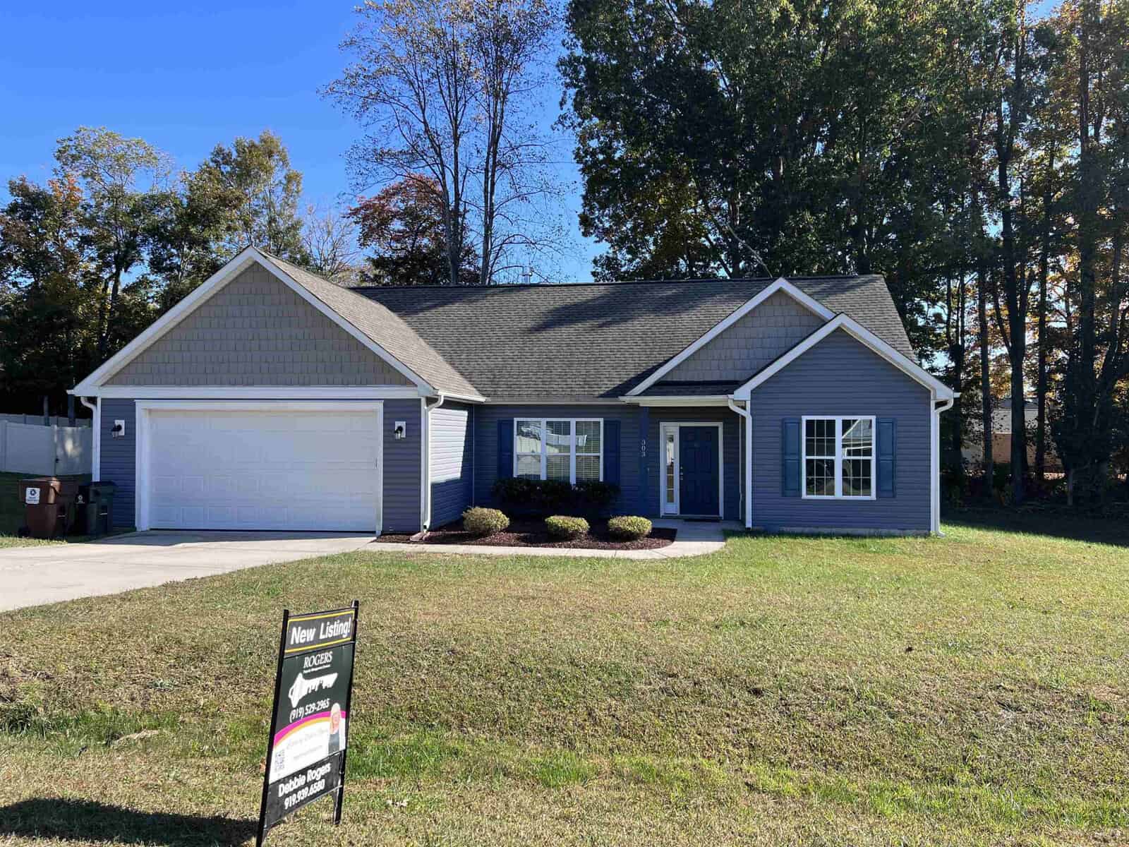 A blue single-story house at 303 Keeneland Drive, Oxford, features a gray roof, two-car garage, and a well-kept lawn. It is surrounded by tall trees and boasts a "New Listing" real estate sign in the front yard.